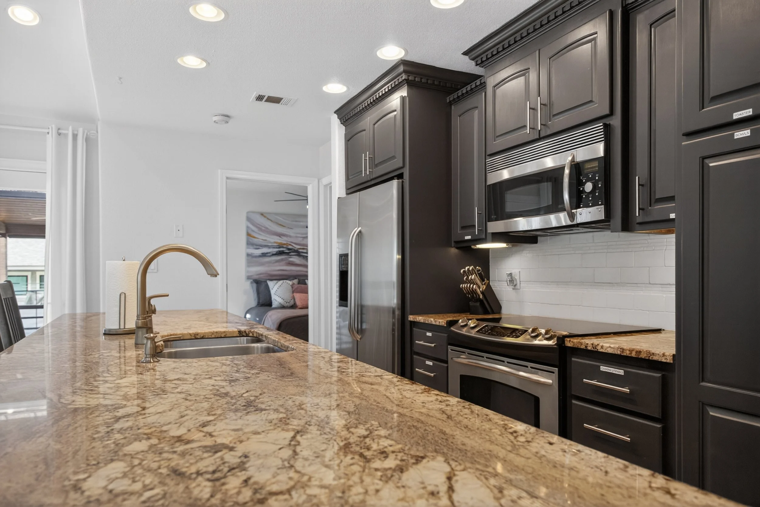 Modern kitchen with dark cabinets, stainless steel microwave and refrigerator, granite countertops, and a view into a bedroom with a bed and abstract artwork.