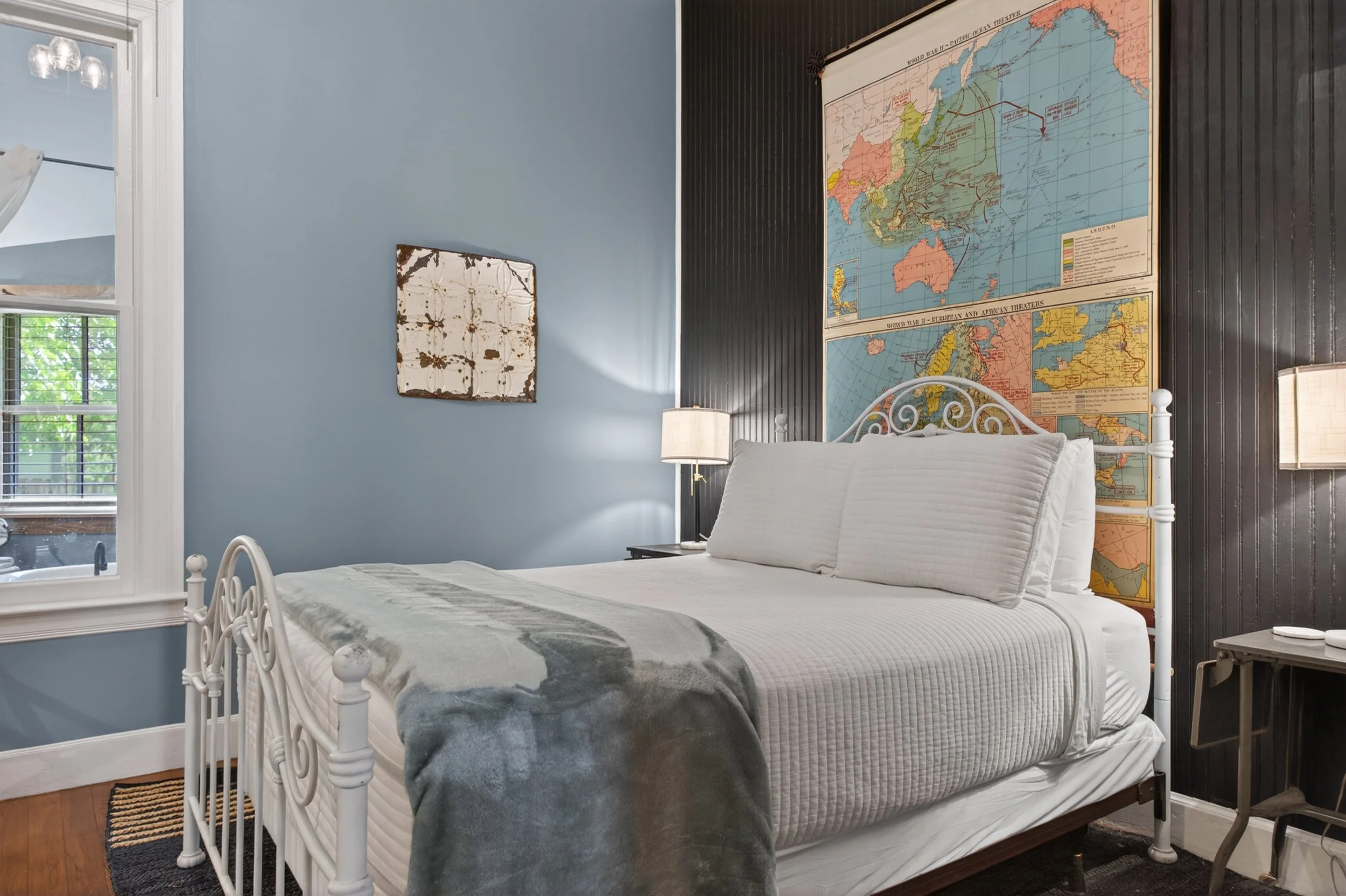 A bedroom with a white metal bed frame, a quilted headboard, and white bedding including pillows and a blanket. There are two lamps on either side of the bed, a wall map of the world behind the headboard, a small window with blinds to the left, and a