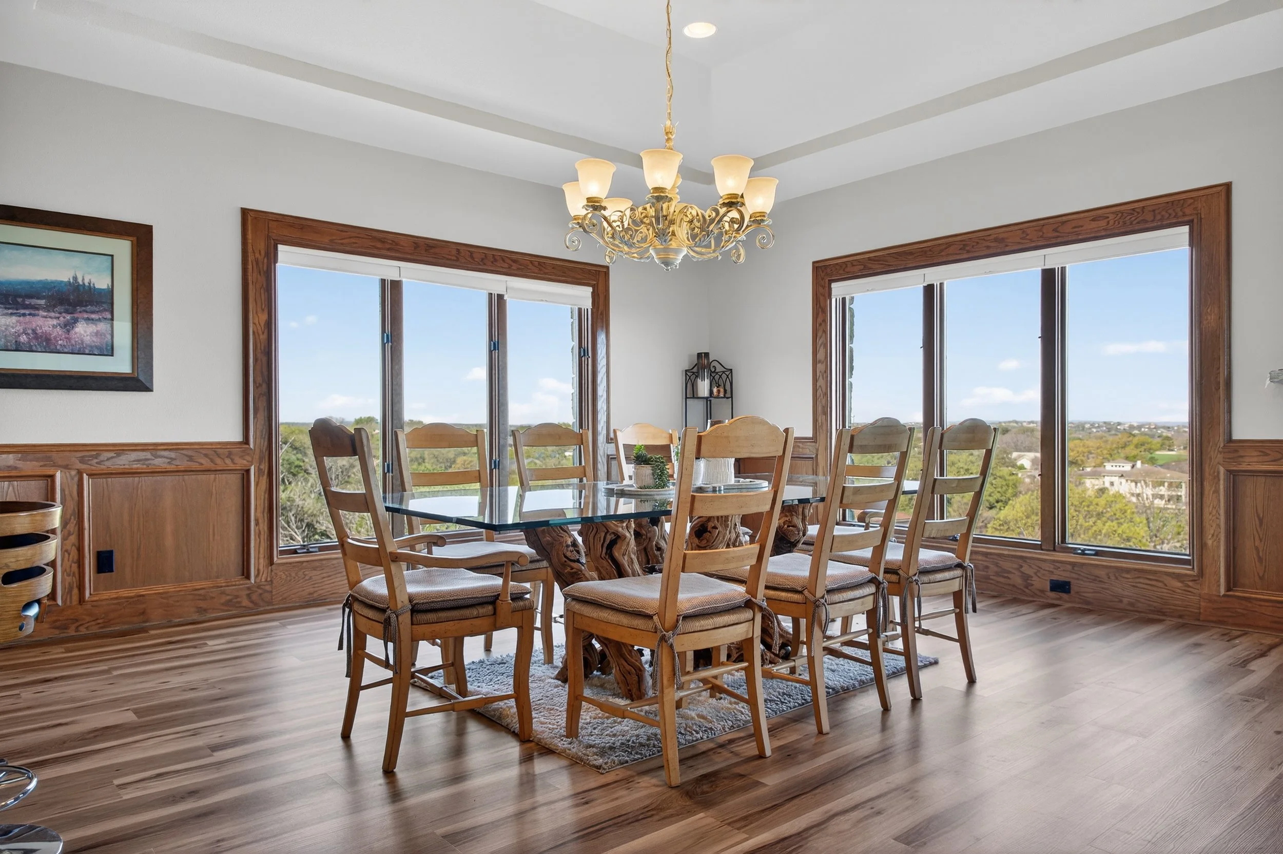 A bright dining room with a wooden dining table and multiple wooden chairs, large windows with views of trees and sky, a chandelier hanging from the ceiling, a decorative shelf in the corner, and framed artwork on the walls.