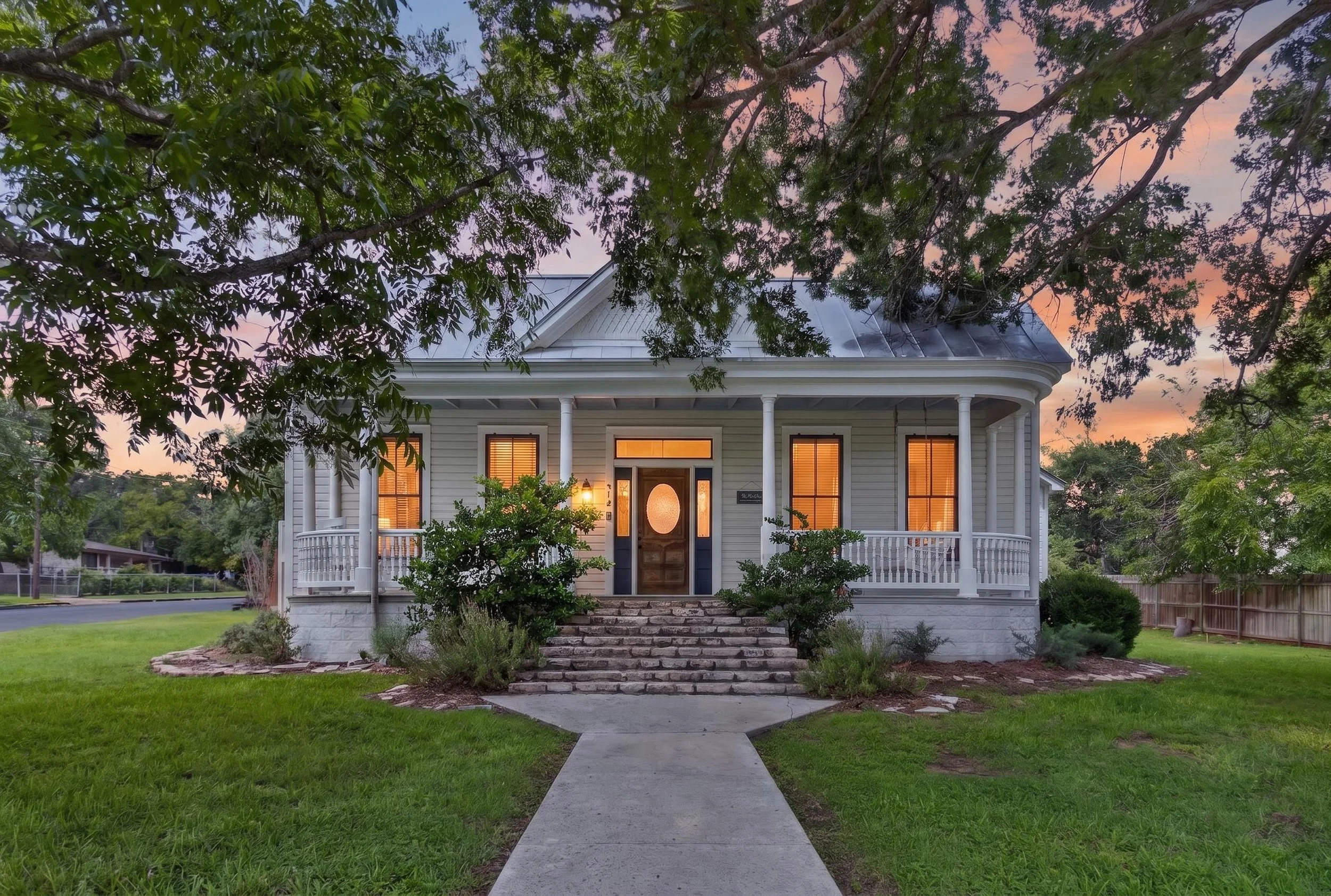 A large, white, two-story house with a front porch, columns, and brick stairs, surrounded by greenery and trees, during sunset.