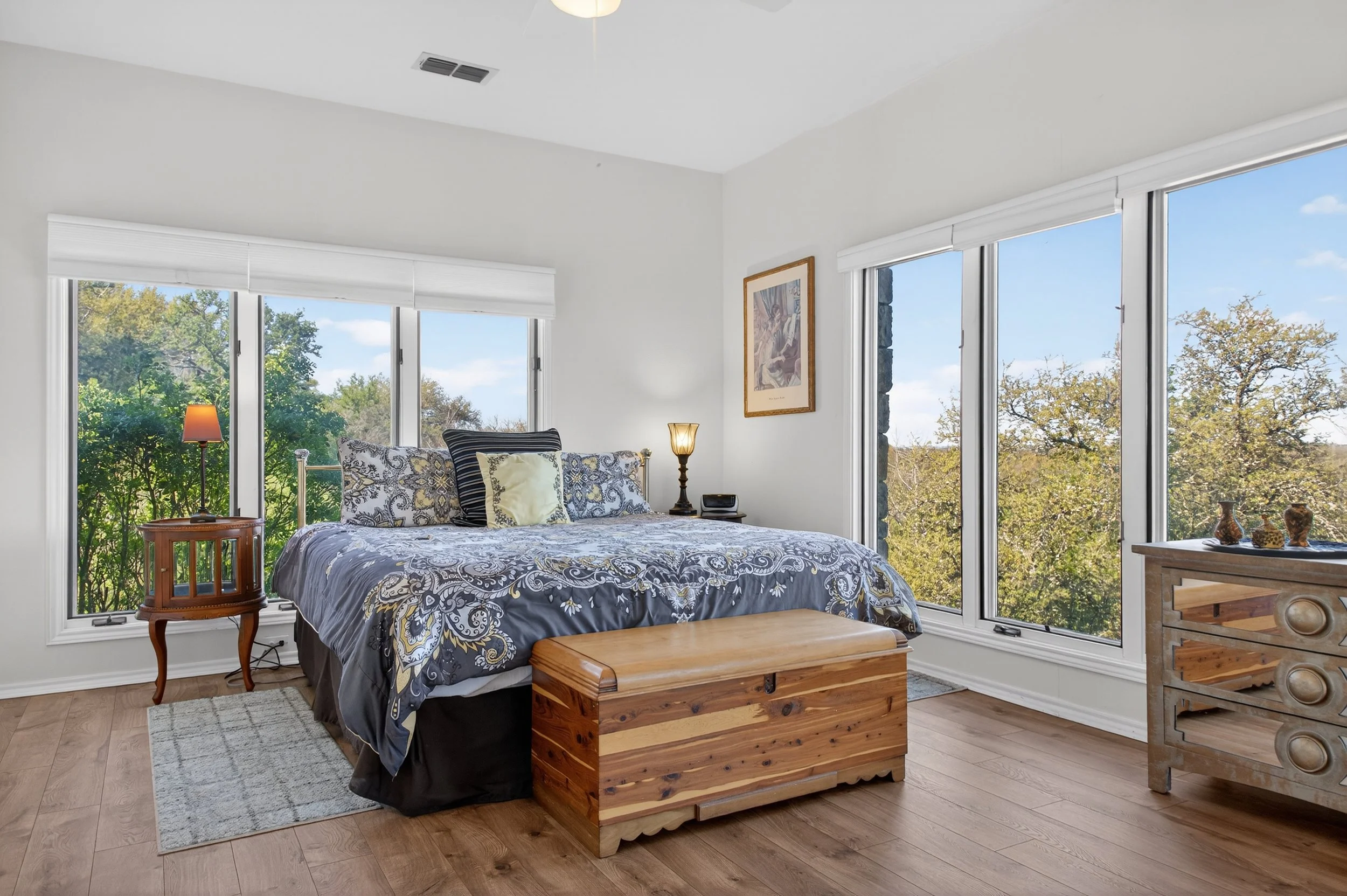 A bedroom with large windows of trees outside, a bed with patterned bedding and pillows, two nightstands with lamps, a wooden chest at the foot of the bed, and a wooden dresser with decorative items.