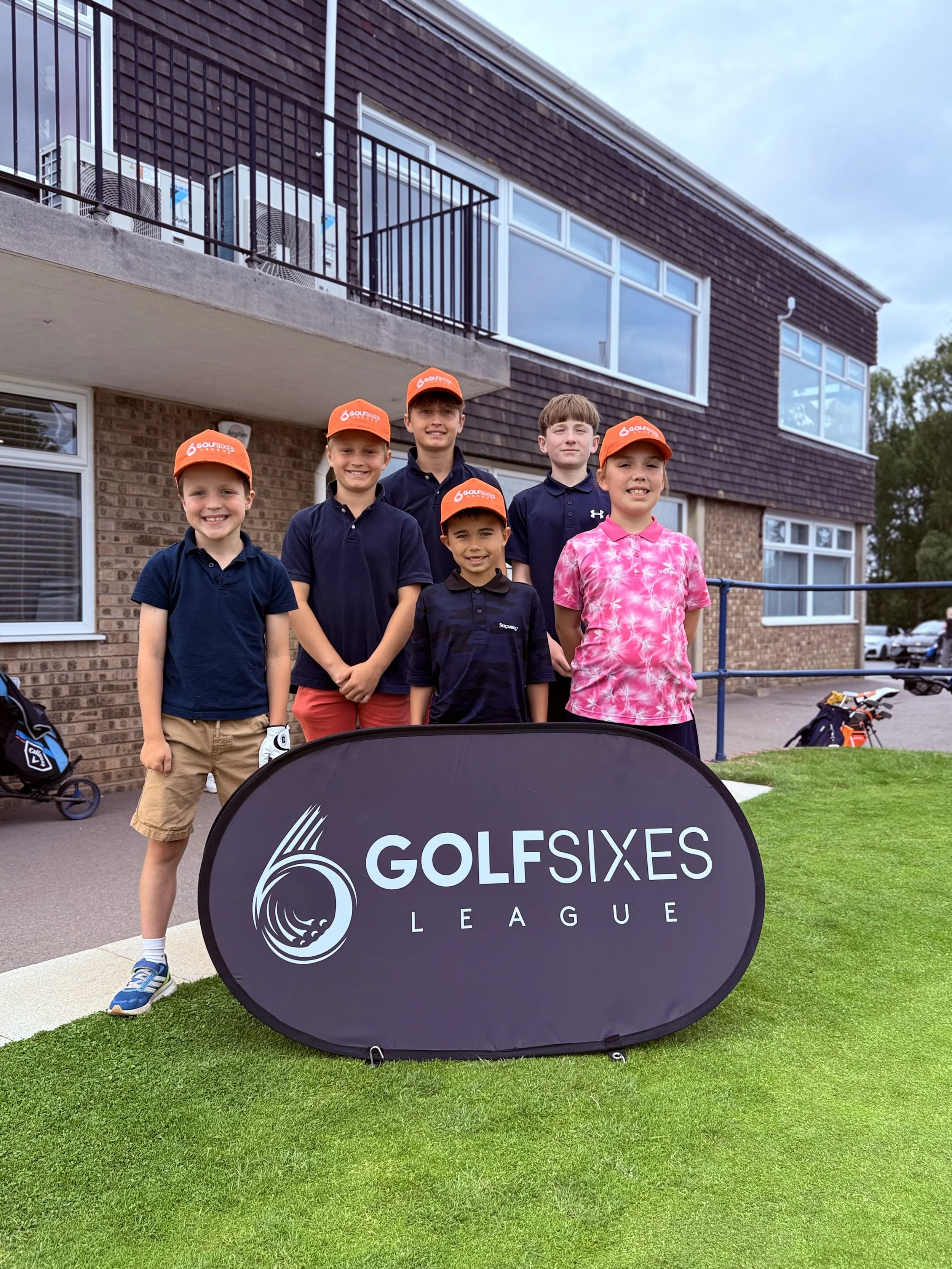 Four boys standing outdoors in front of a building, wearing golf hats and shirts, with a black sign that reads "GOLFSIXES LEAGUE" in the foreground.