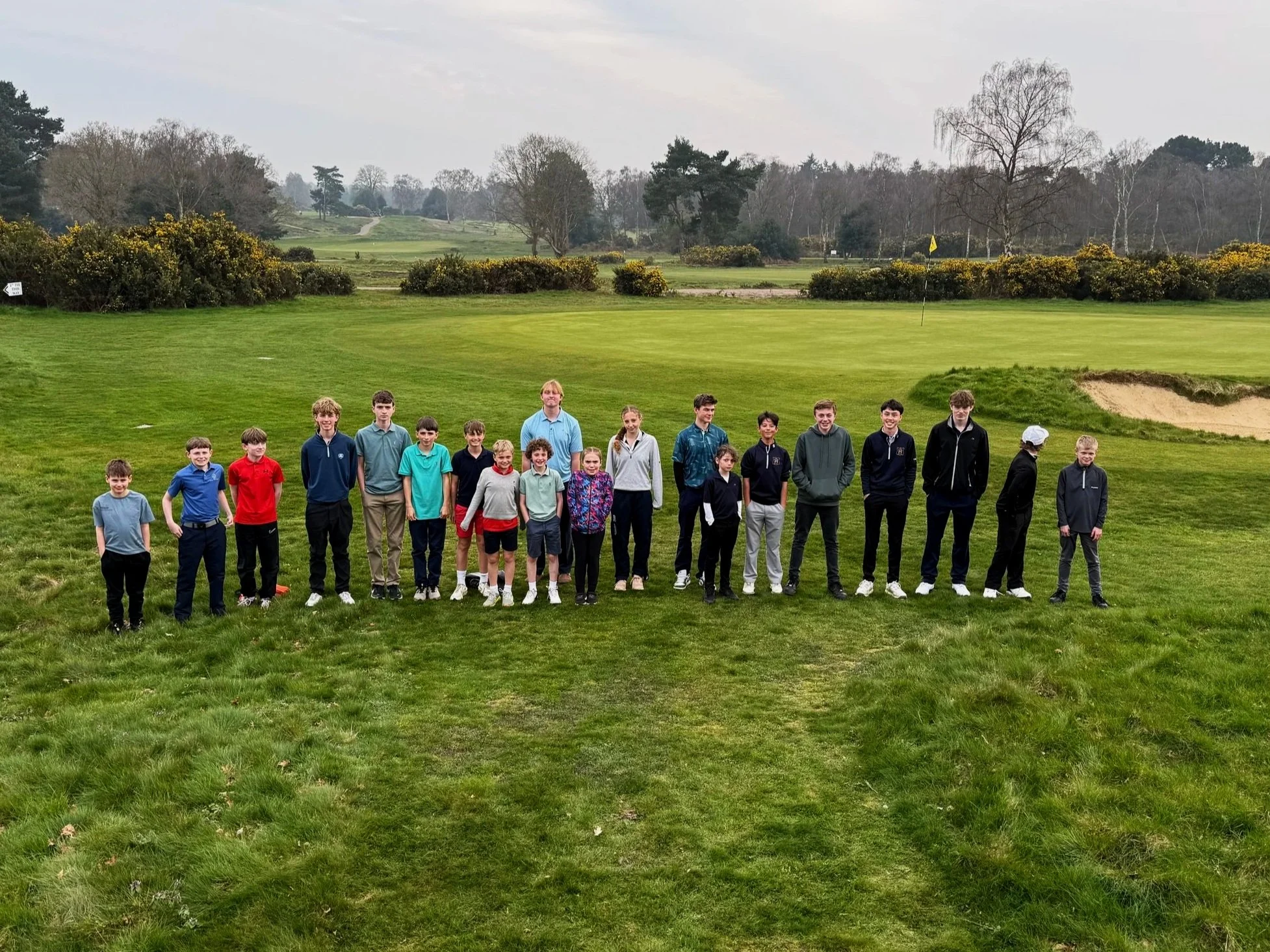 A group of children and teenagers standing on a golf course near a putting green, with trees and hills in the background.