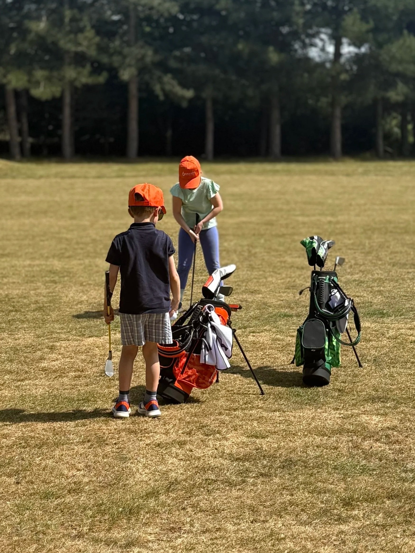 Two children in golf attire on a golf course, one girl and one boy, standing with golf clubs and golf bags.