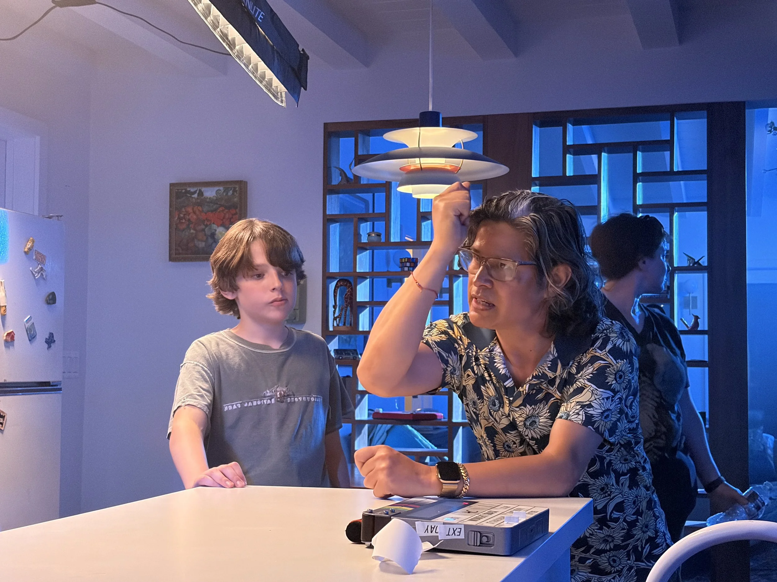 A woman and a young boy are engaged in an arm wrestling match at a table. The woman is wearing glasses, a floral shirt, and a smartwatch, while the boy has medium-length brown hair and is wearing a gray t-shirt. The background features blue lighting, a wooden shelving unit with decorative items, and a framed painting on the wall.