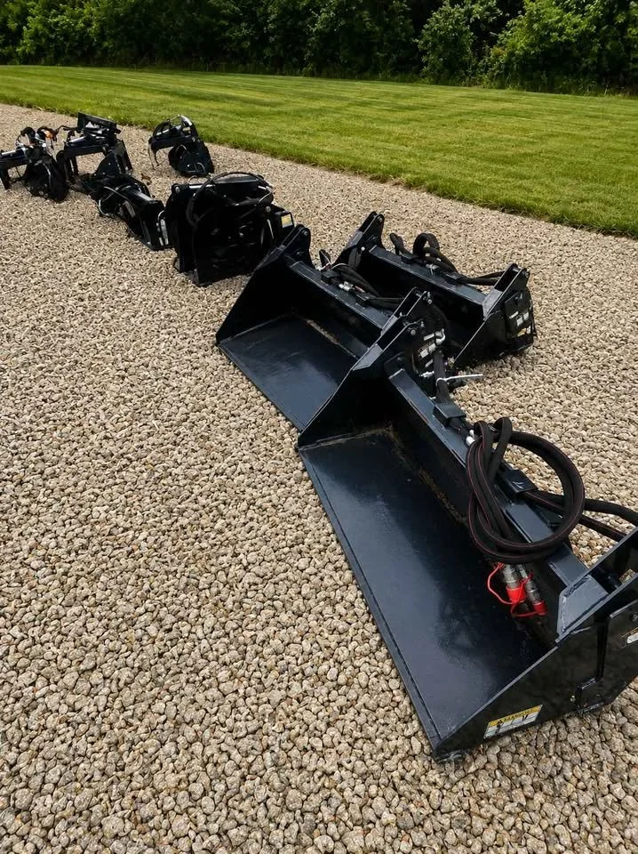 A row of black skid steer attachments arranged on a gravel surface with grass and trees in the background.