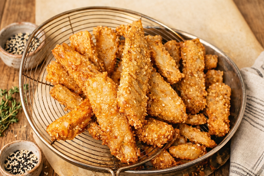 A wire basket filled with crispy, breaded chicken tenders on a wooden table with small bowls of black and white sesame seeds nearby.