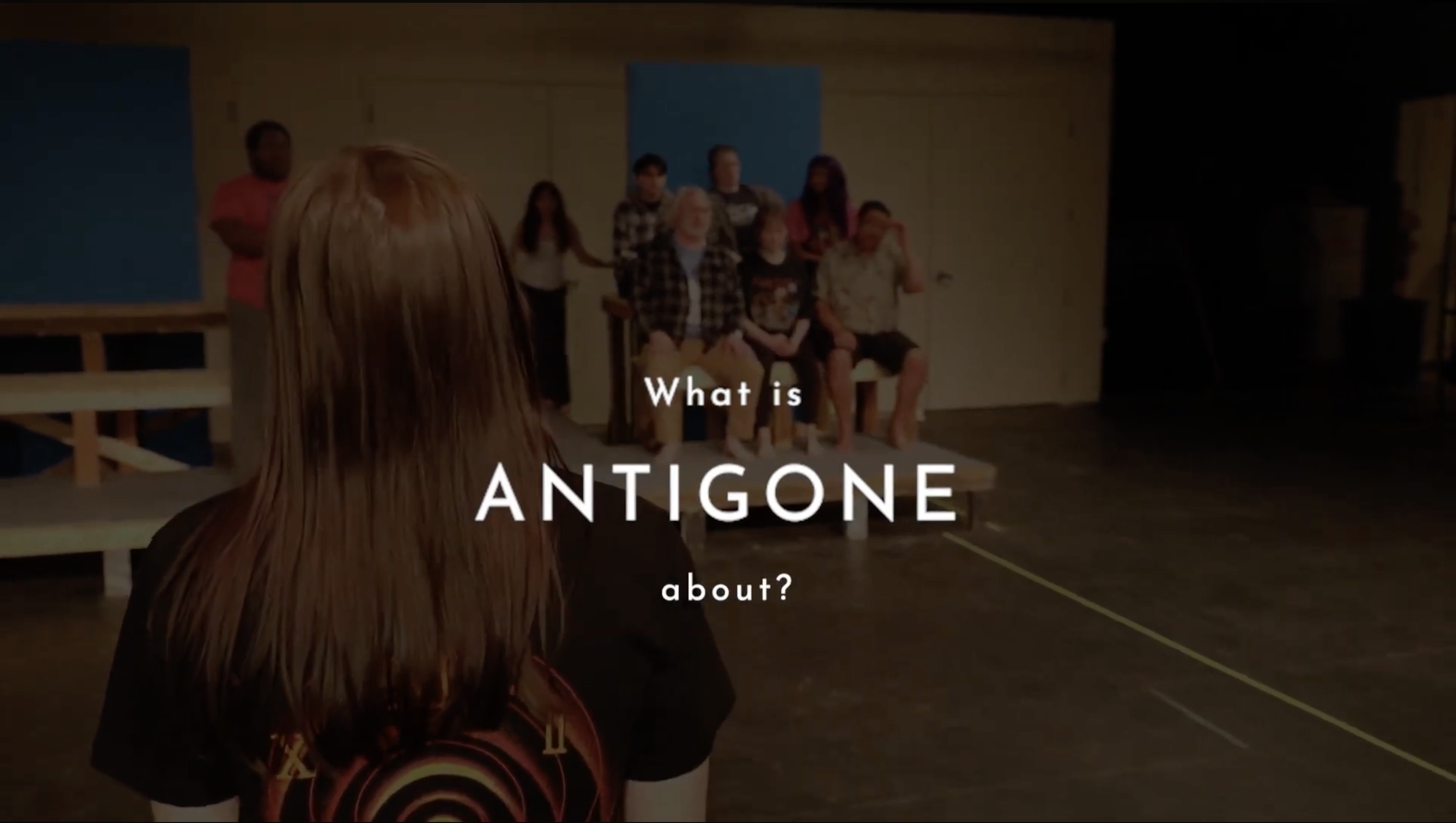Words across actors in the background. The words read: "What is Antigone about?"