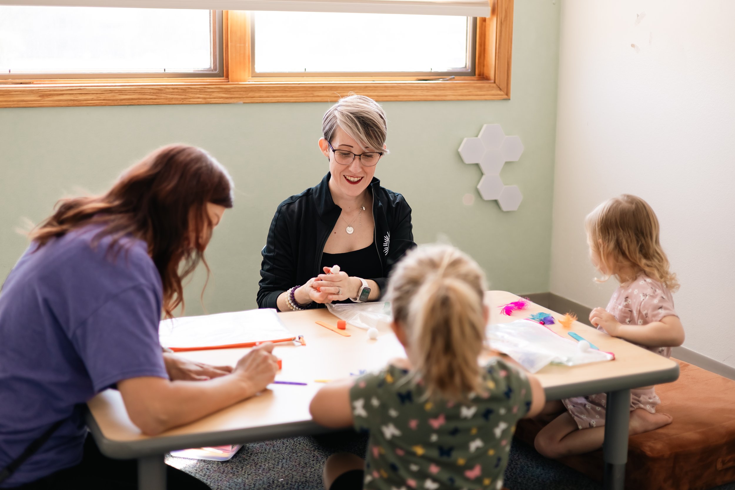 A group of women and children sitting around a table in a brightly lit room with a window, engaging in arts and crafts activities.