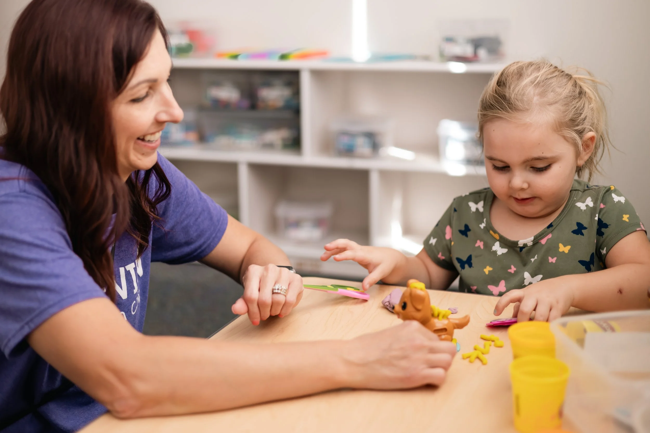 A woman with long dark hair smiling and a young girl with blonde hair playing with colorful clay on a wooden table in a room with shelves of organized craft supplies in the background.