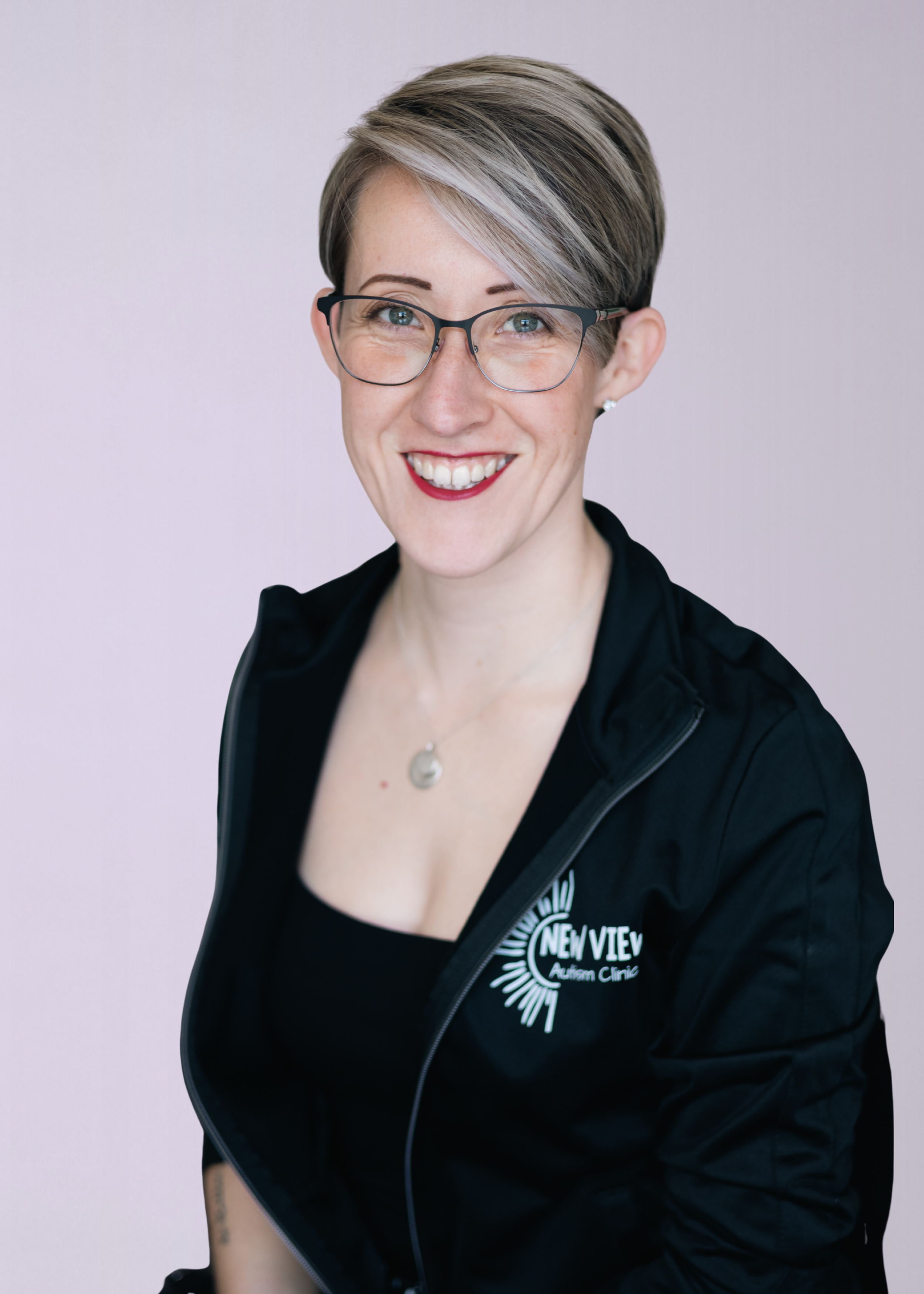 Smiling woman with short, colored hair wearing glasses and a black jacket with the logo 'New View Autism Clinic'.