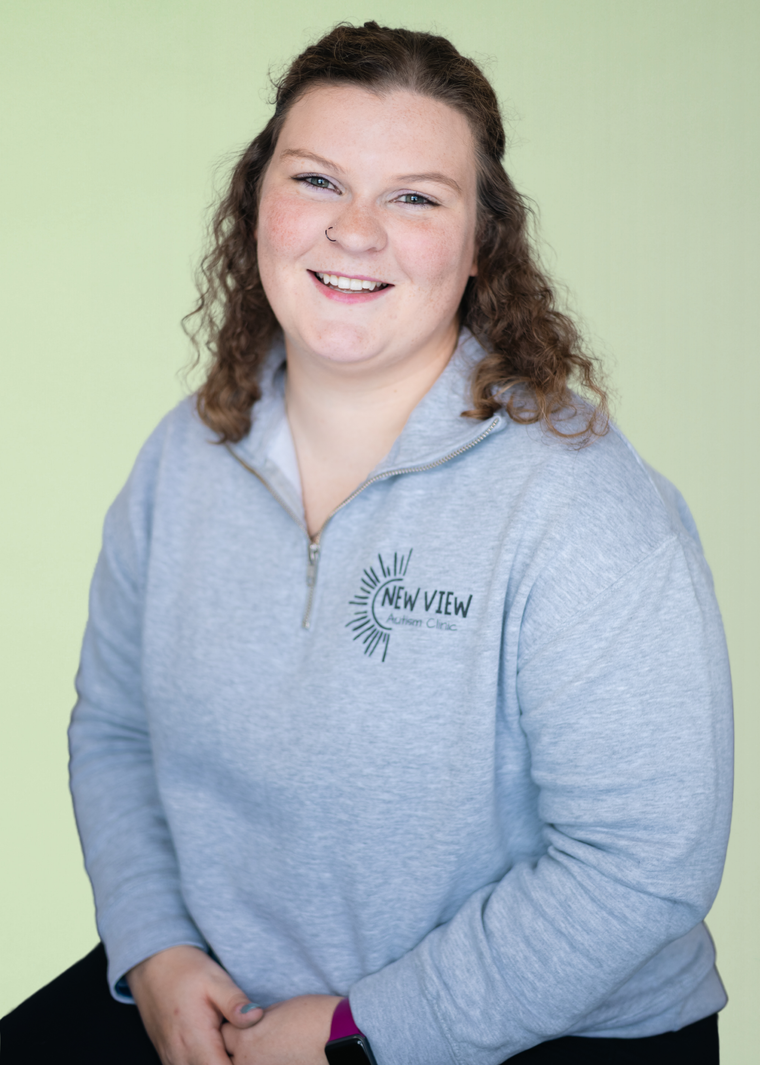 A smiling woman with curly brown hair, wearing a gray quarter-zip pullover with 'New View Autism Clinic' logo, seated against a light green background.