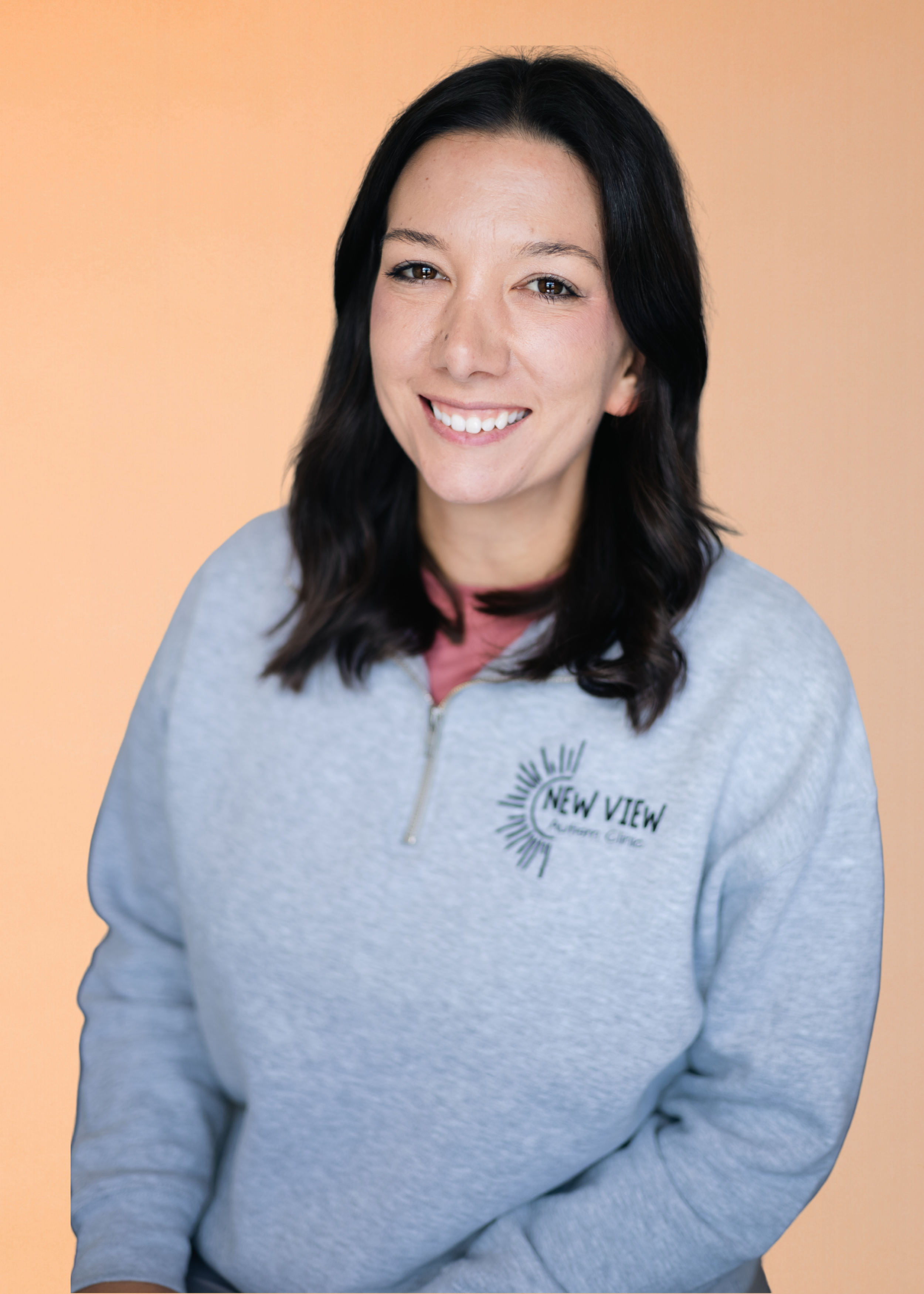 A woman with long dark hair, smiling, wearing a gray sweatshirt with a logo that reads "New View" against an orange background.