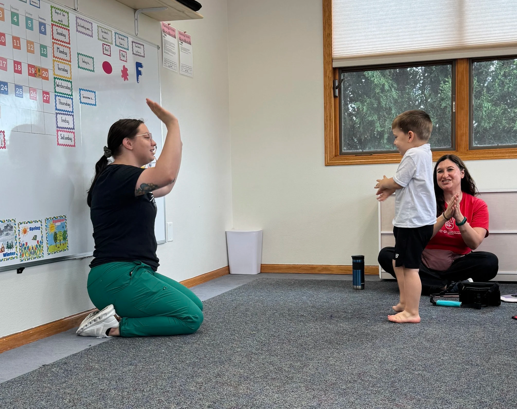 A woman kneeling on a dark gray carpet, raising her hand to high-five a young boy who is standing on the same carpet. Both are smiling and engaged in an activity, possibly a classroom or therapy session. Another woman is sitting nearby, clapping and smiling. Behind them is a window with a view of trees and a whiteboard with colorful calendar and educational posters.