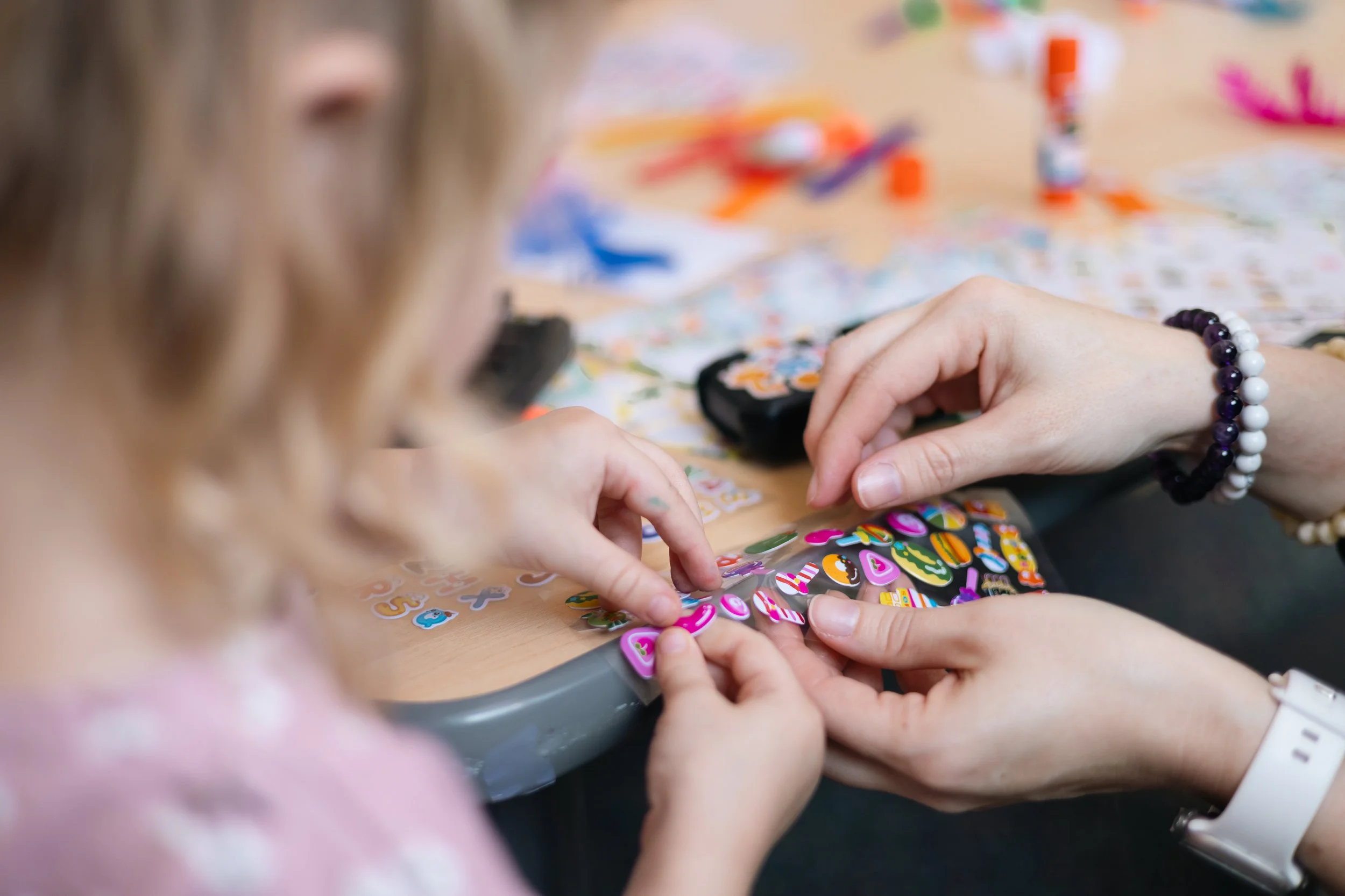 Close-up of children playing a sticker game with colorful stickers and game pieces on a table, with one child's hand reaching for stickers.