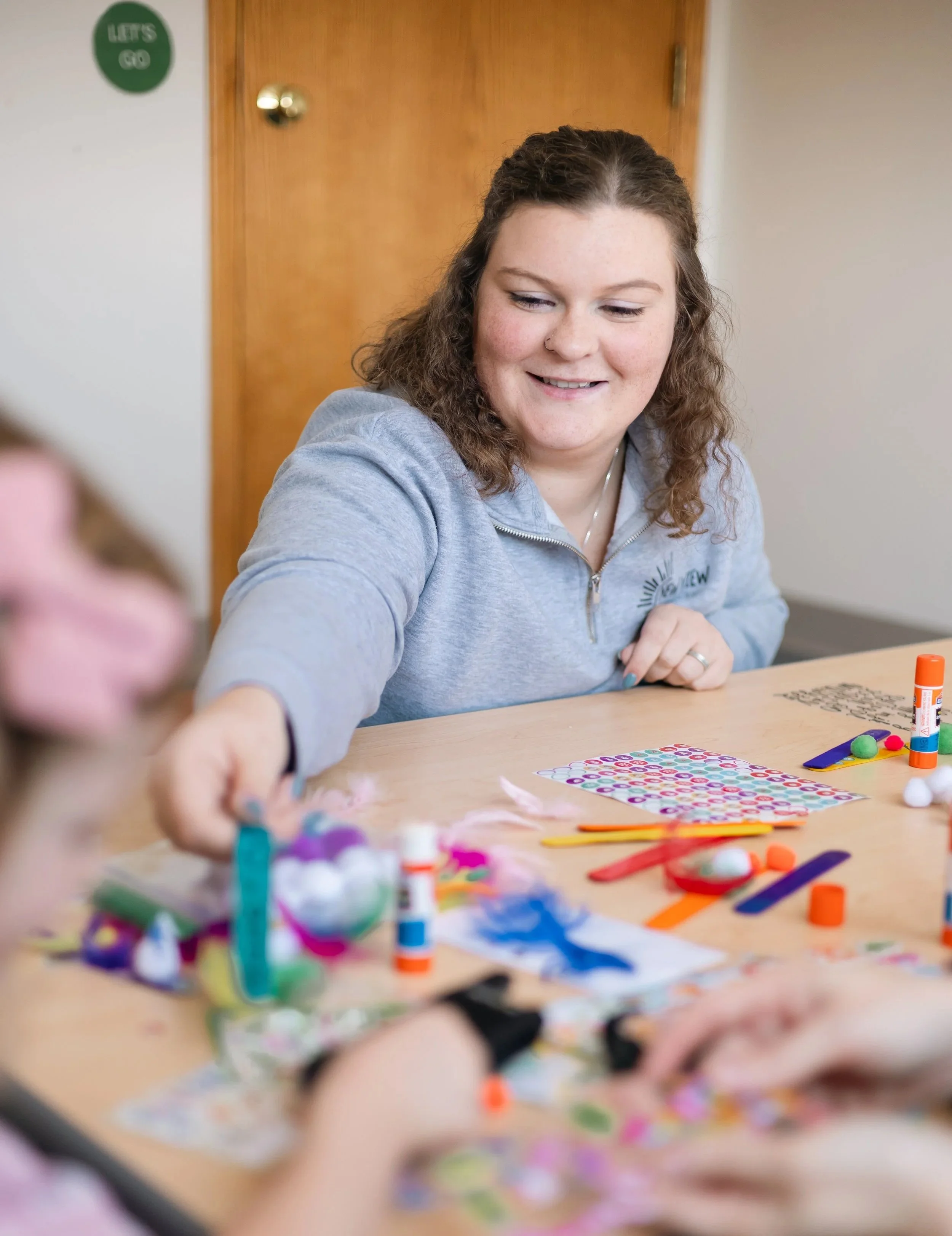 Teenager smiling while working on arts and crafts project with colorful paper, glue, and decorations on a table.