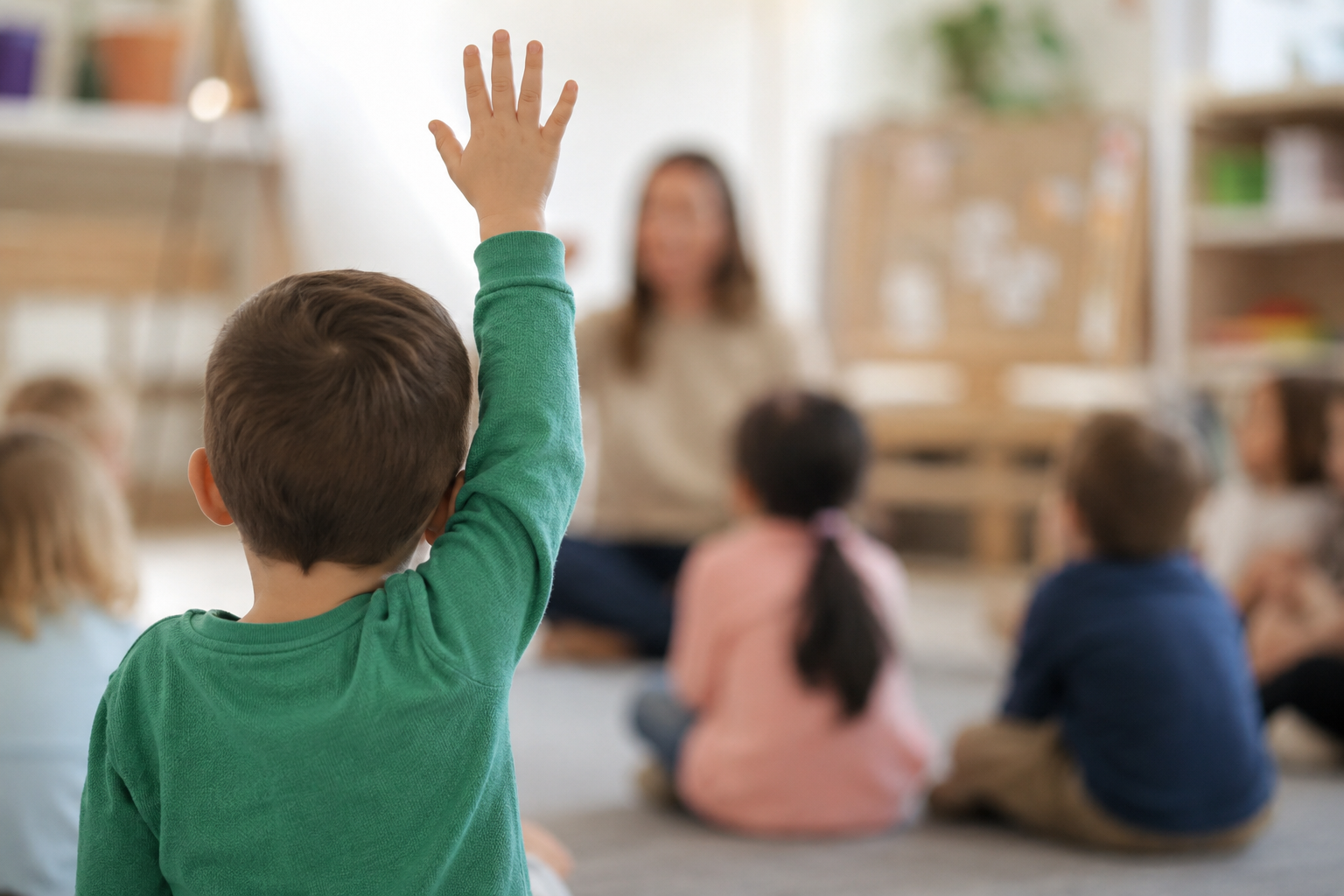 A young boy with brown hair in a green shirt raising his hand in a classroom filled with children sitting on the floor. A woman, likely a teacher, is sitting in front of them. The classroom has shelves and boxes in the background.