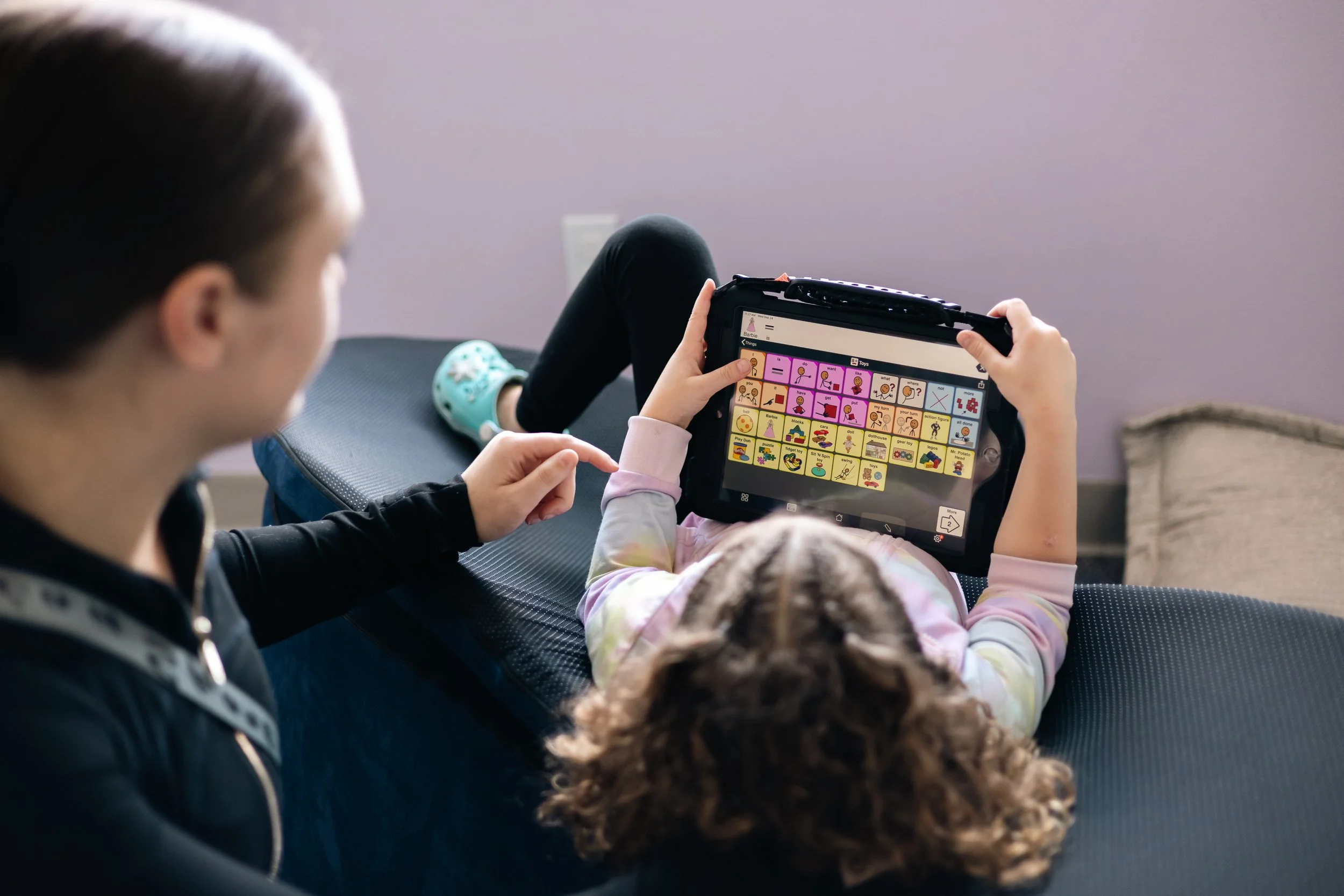 A young girl with curly hair is using a communication device with pictures and symbols, possibly for speech assistance, while sitting on a couch.