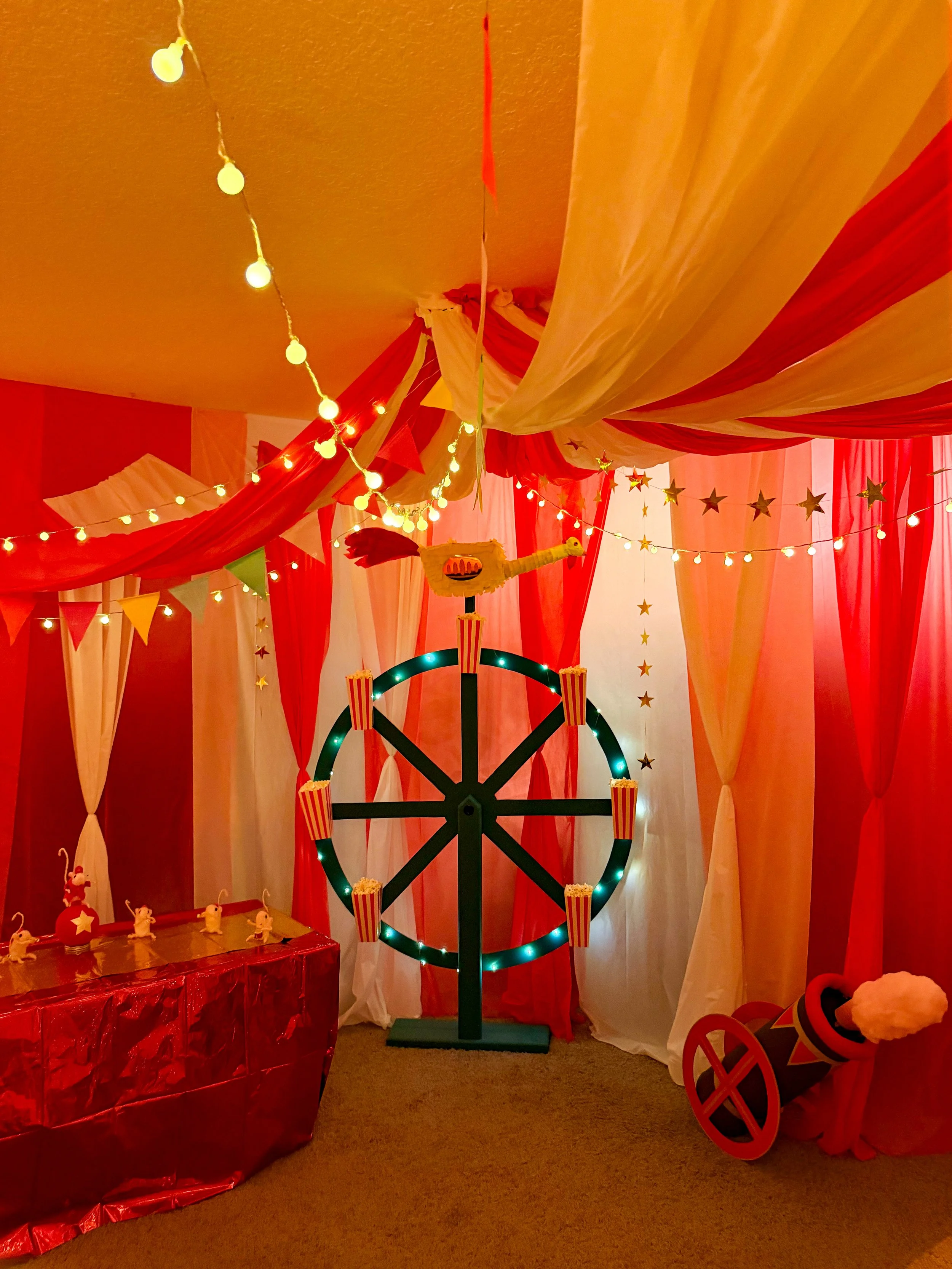 Red-and-white circus-themed party room with string lights, star decorations, popcorn boxes, a cannon, and draped fabric.