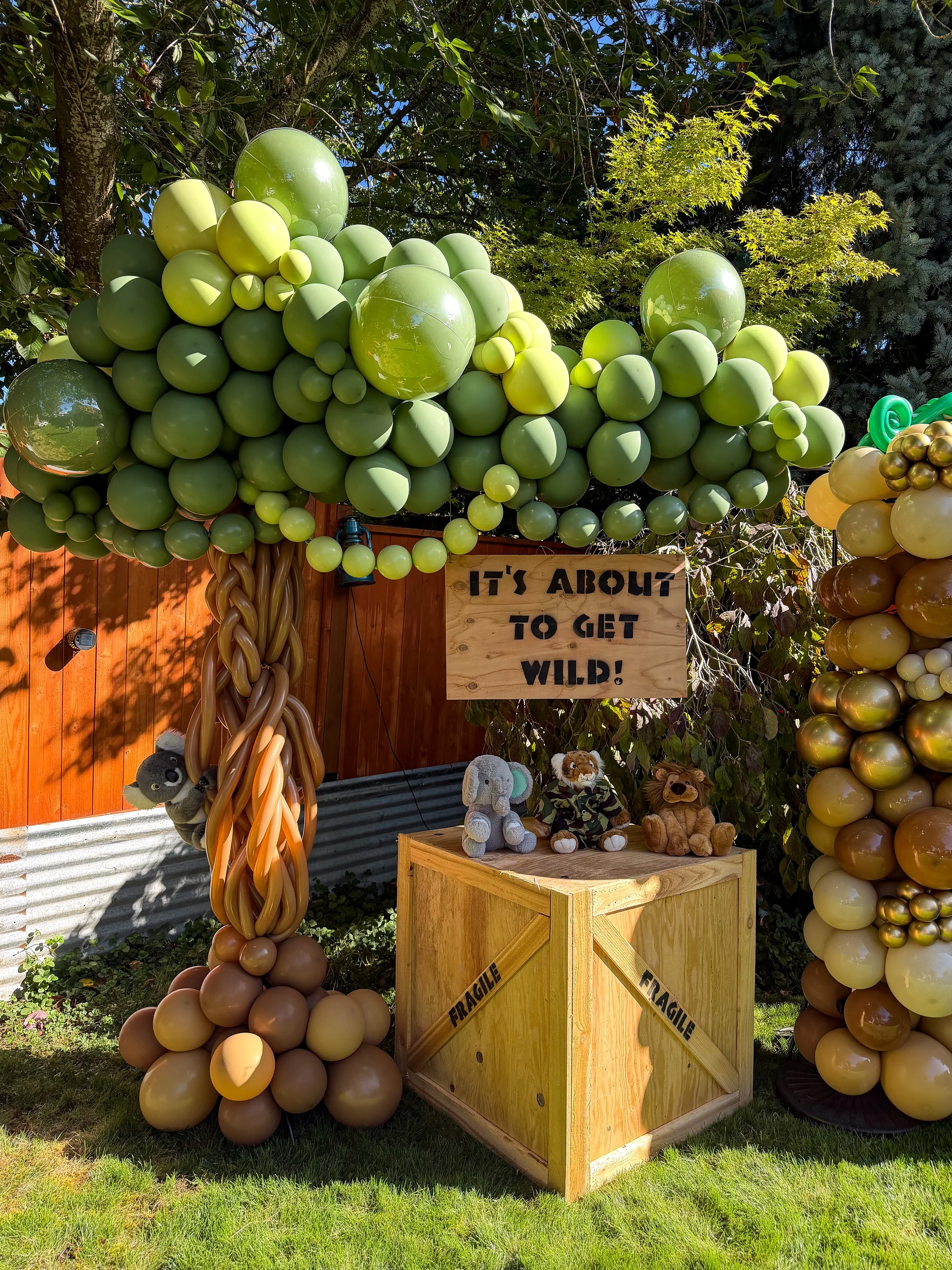 Animal-themed balloon display with green balloon arch, woodland stuffed animals, wooden crates, and a sign reading “It’s About to Get Wild!”
