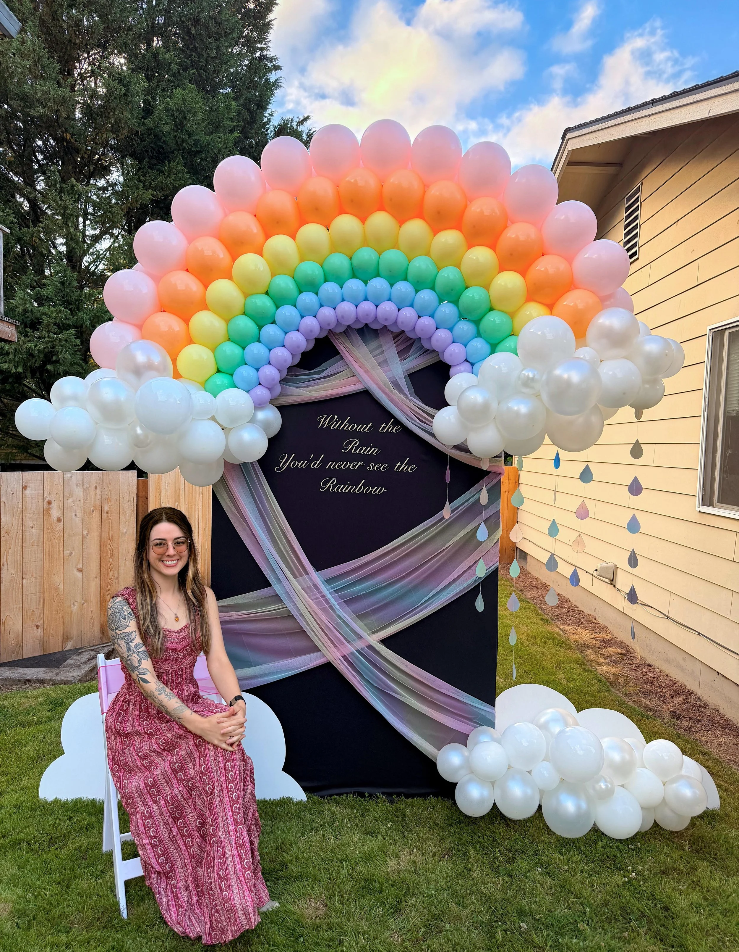 Woman in a pink dress seated beside a colorful rainbow balloon arch and backdrop decorated with white balloon clouds outdoors.