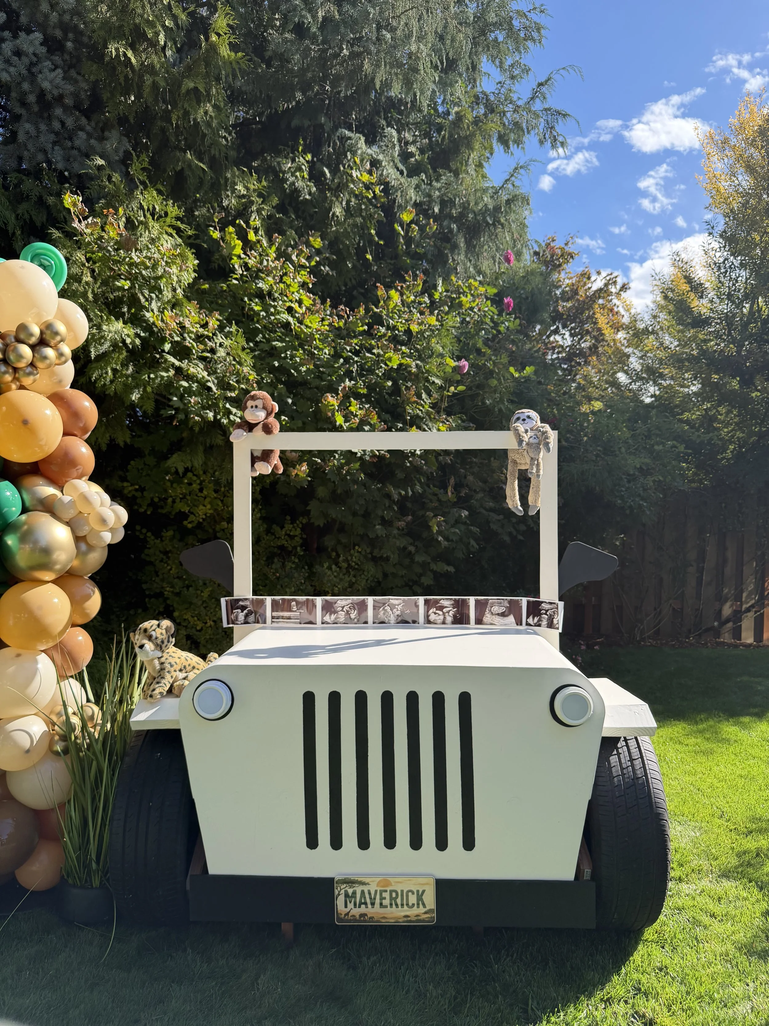 Outdoor jungle-themed baby shower display with a white Jeep prop, balloon arch, stuffed monkeys, ultrasound photo strip, and lush garden backdrop.