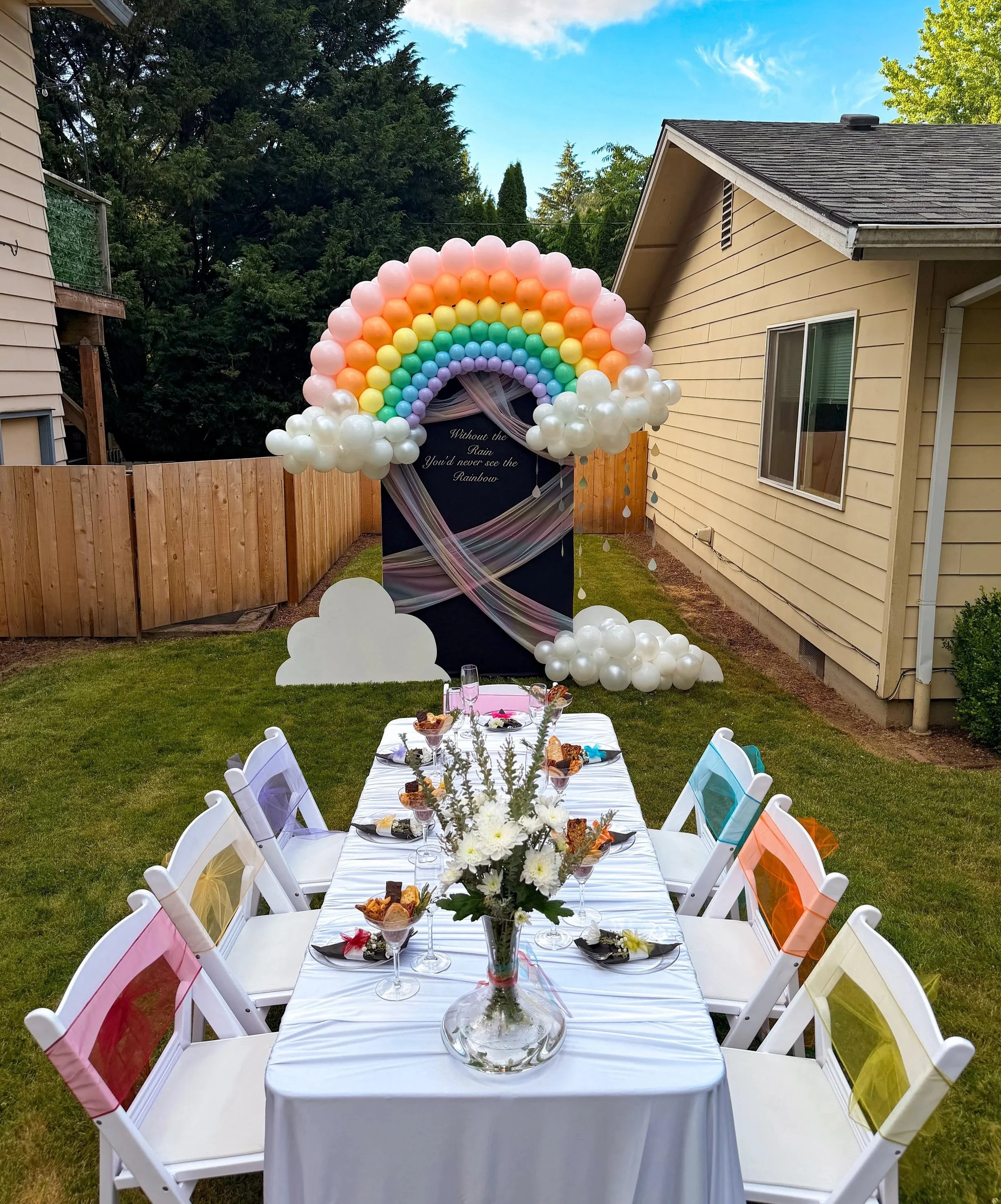 Outdoor celebration with a rainbow balloon arch, decorated dining table, white folding chairs, and floral centerpiece in a backyard.
