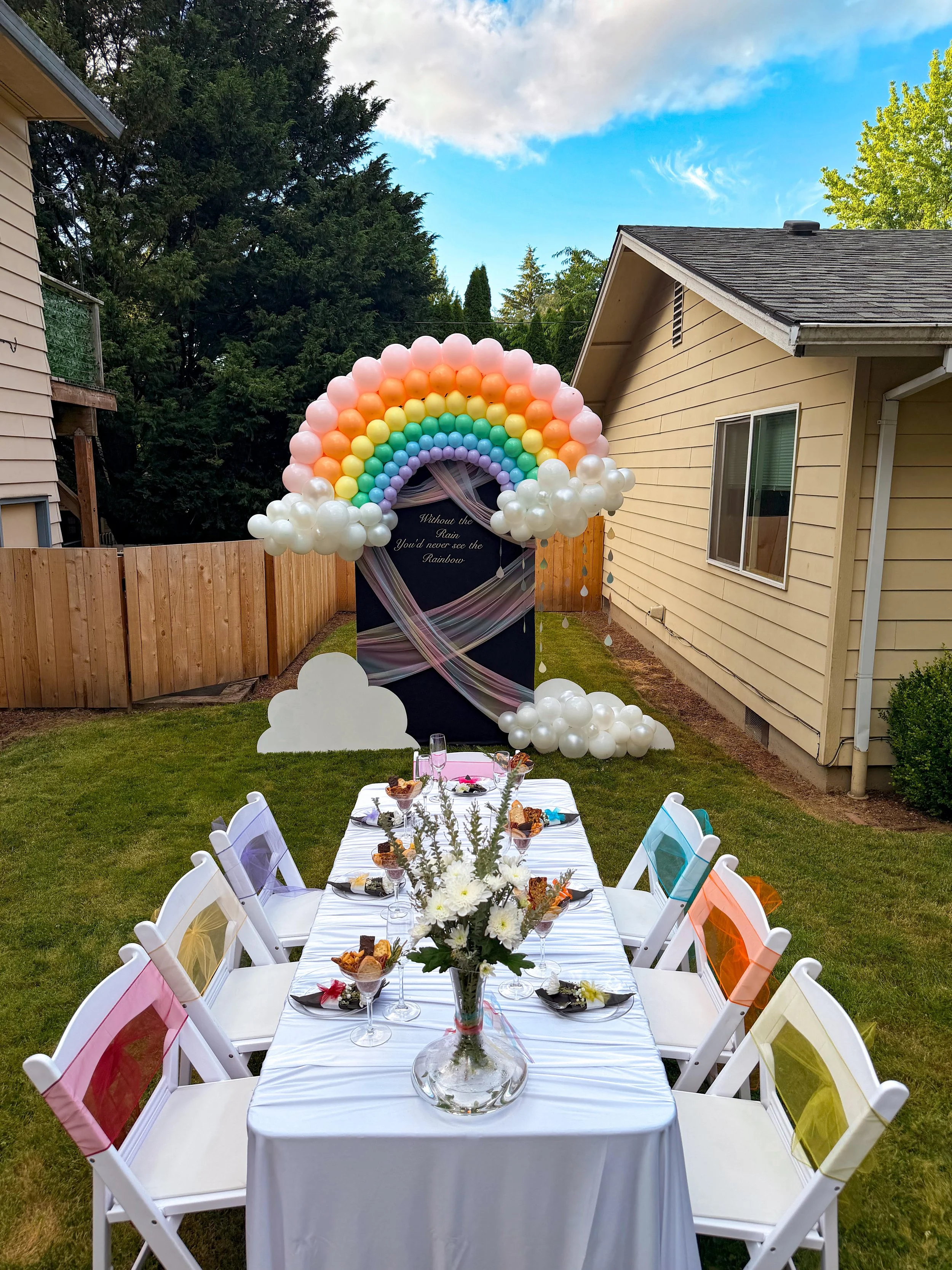 Outdoor celebration setup with a rainbow balloon arch, white balloon clouds, decorated dining table, and colorful chairs in a backyard.