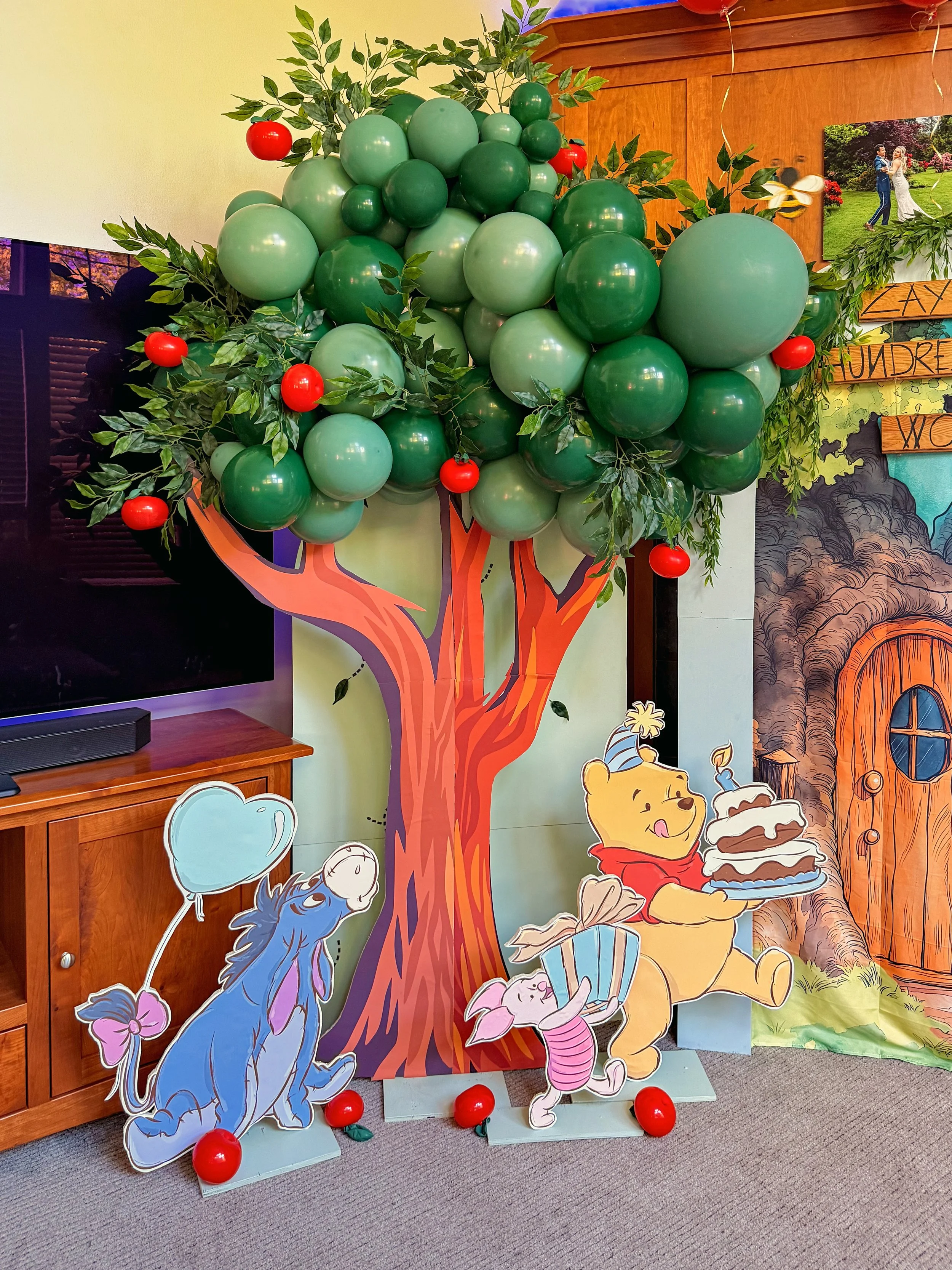 Winnie-the-Pooh-themed party display with a large green balloon tree, leafy branches, red balloons, and cutouts of Eeyore, Piglet, and Pooh holding a cake.