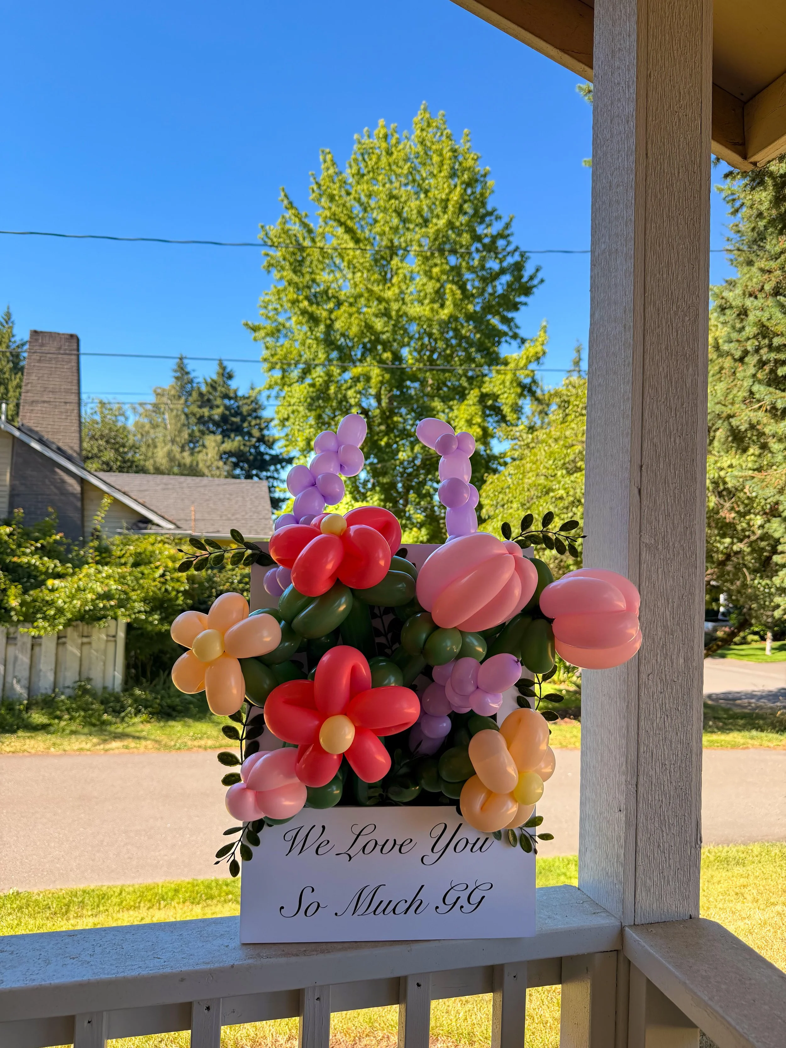 Colorful balloon flower arrangement in a planter with a sign reading “We Love You So Much GG,” displayed on a porch.