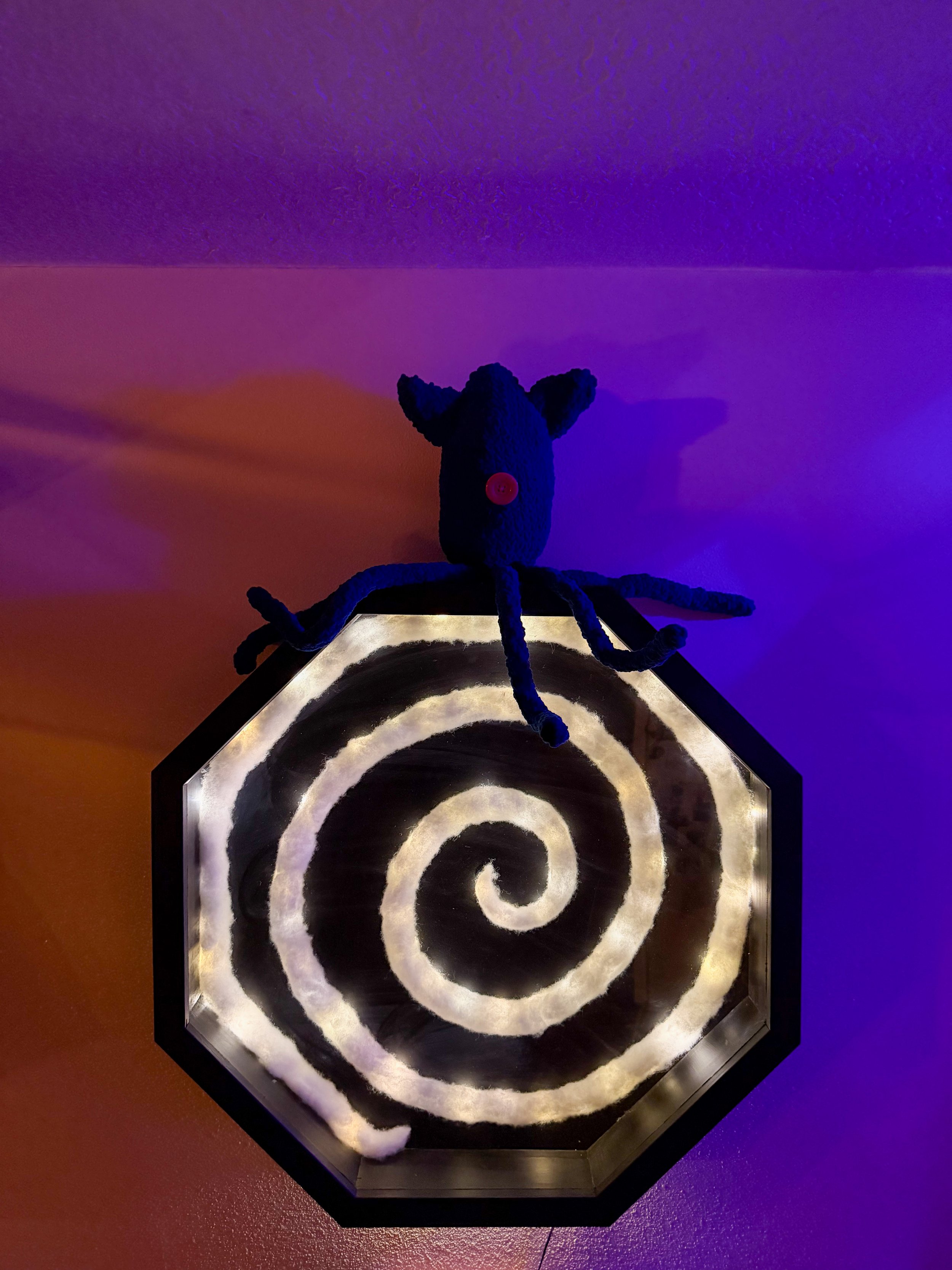 Black plush spider with a red button eye on a spiral-patterned pedestal under purple and orange lighting