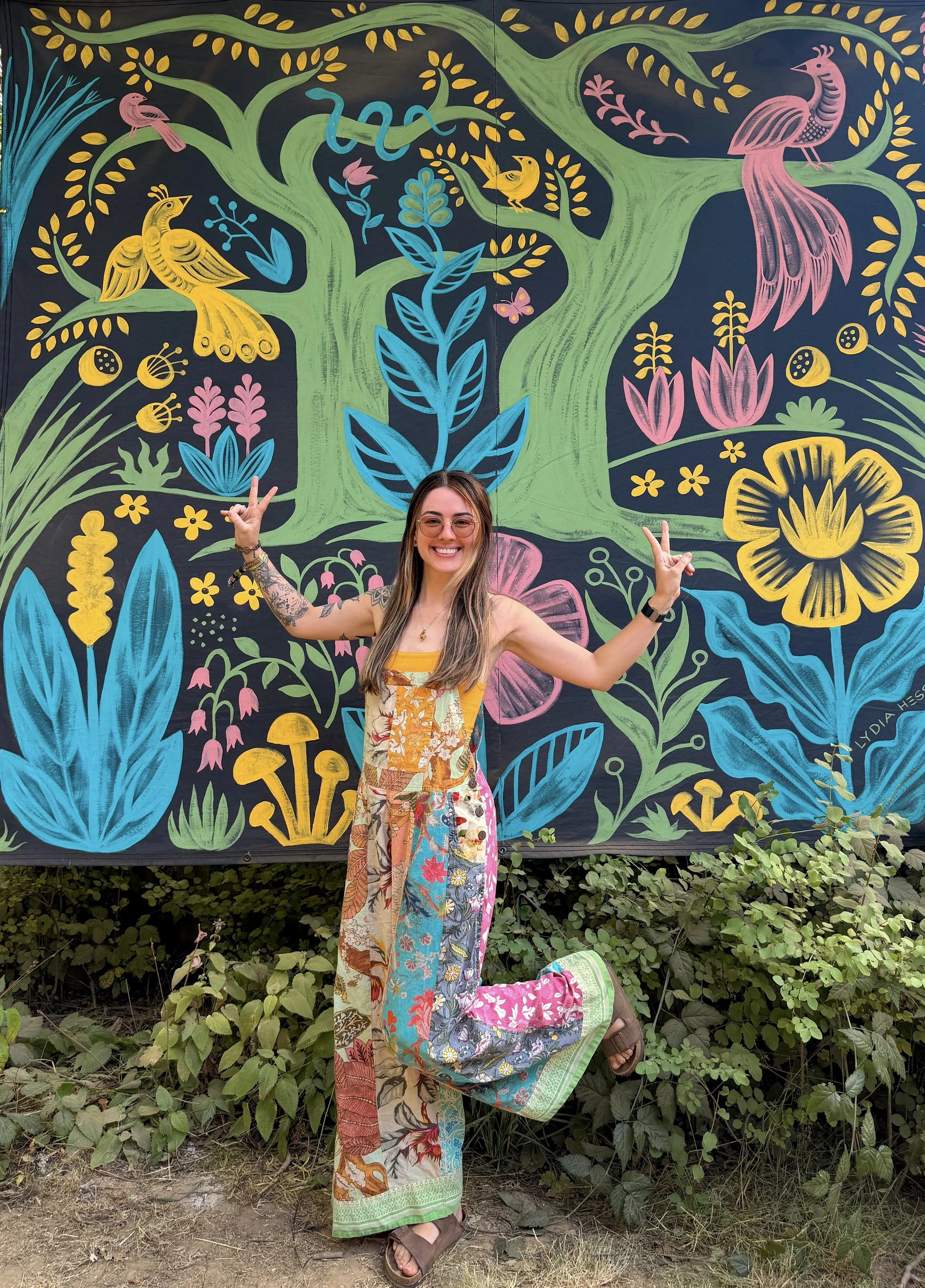 Smiling woman in a colorful dress poses in front of a vibrant painted mural featuring birds, flowers, foliage, and a snake.