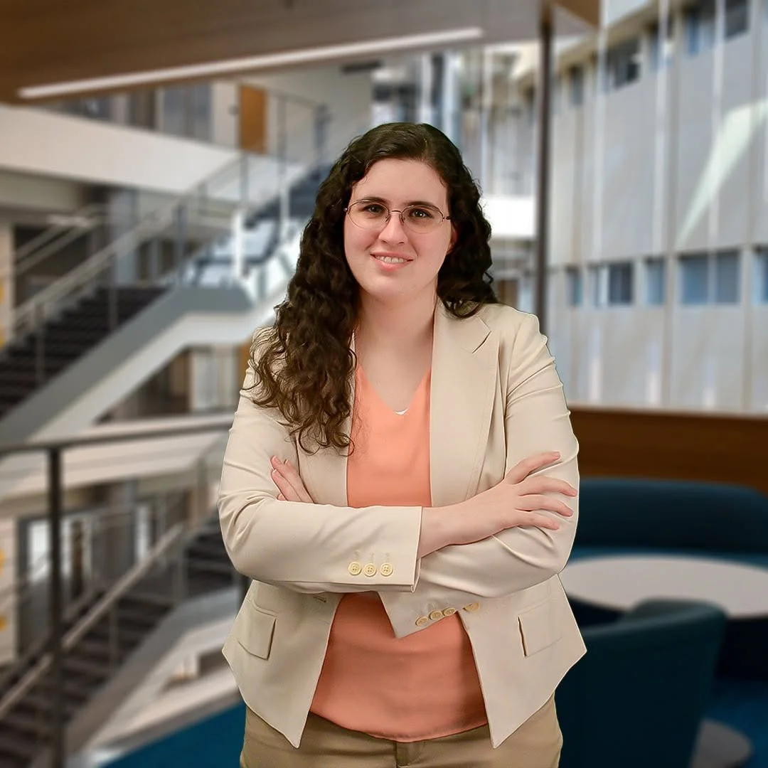 A woman with curly dark hair, glasses, wearing a beige blazer over a peach top, standing with arms crossed in a modern office building.