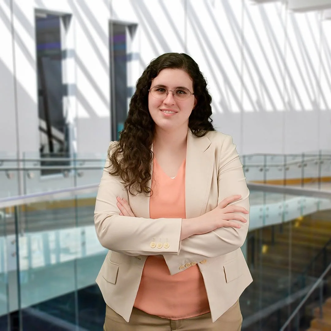 A woman with curly brown hair, glasses, wearing a beige blazer and peach-colored top, standing with crossed arms inside a modern building with architectural shadows on the wall.
