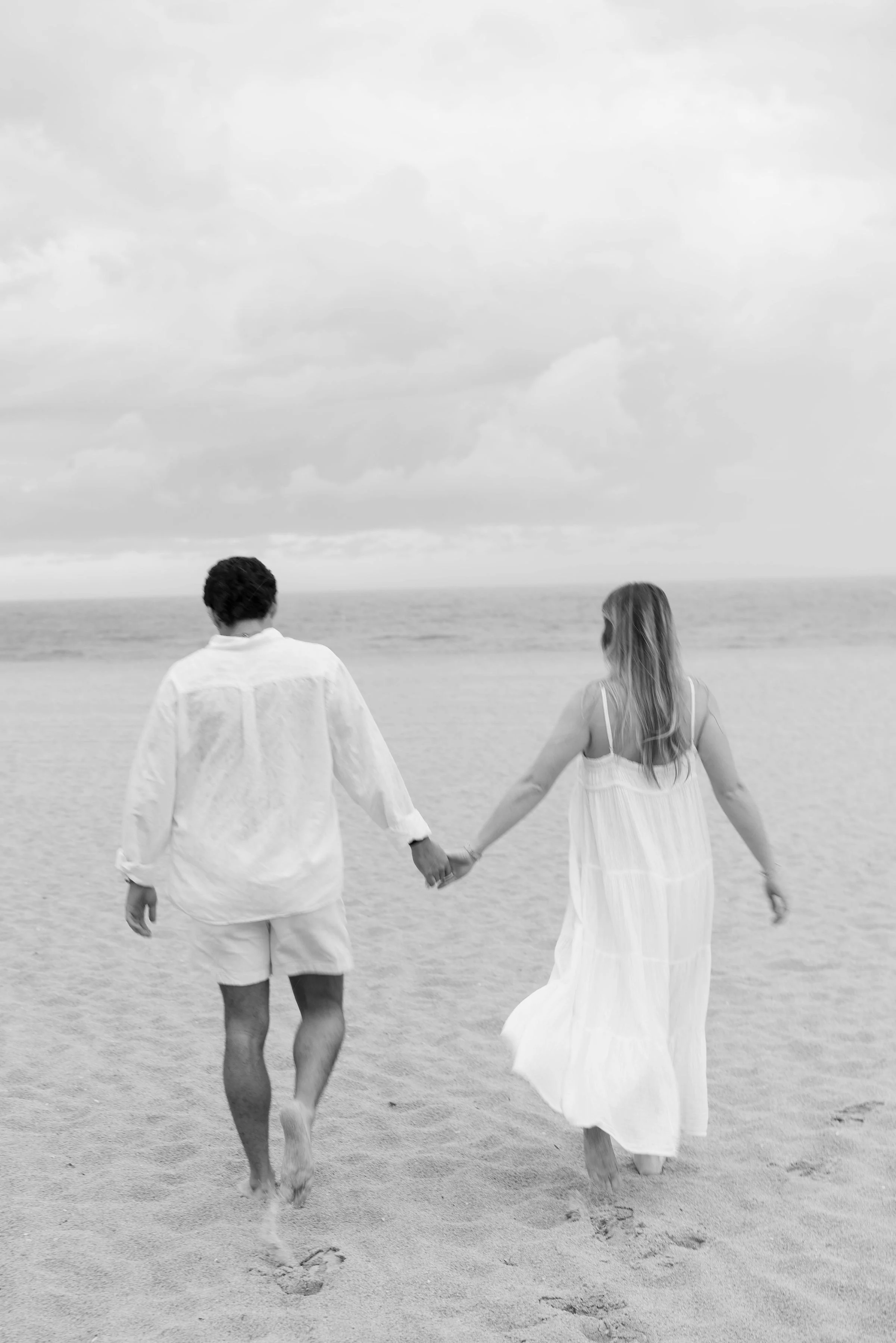 A couple holding hands walking on the beach in white clothing, with the ocean and cloudy sky in the background.