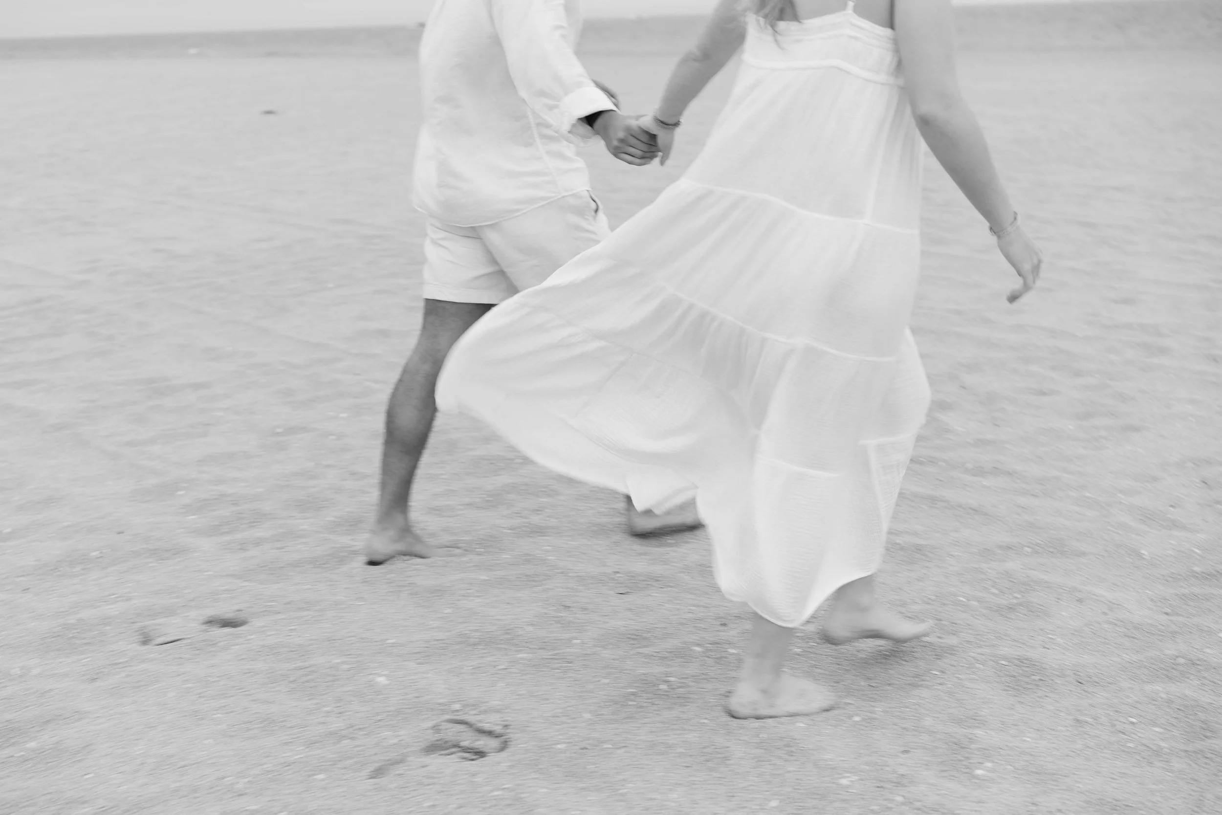 People holding hands and walking along a beach, wearing light clothing, with waves visible in the background.