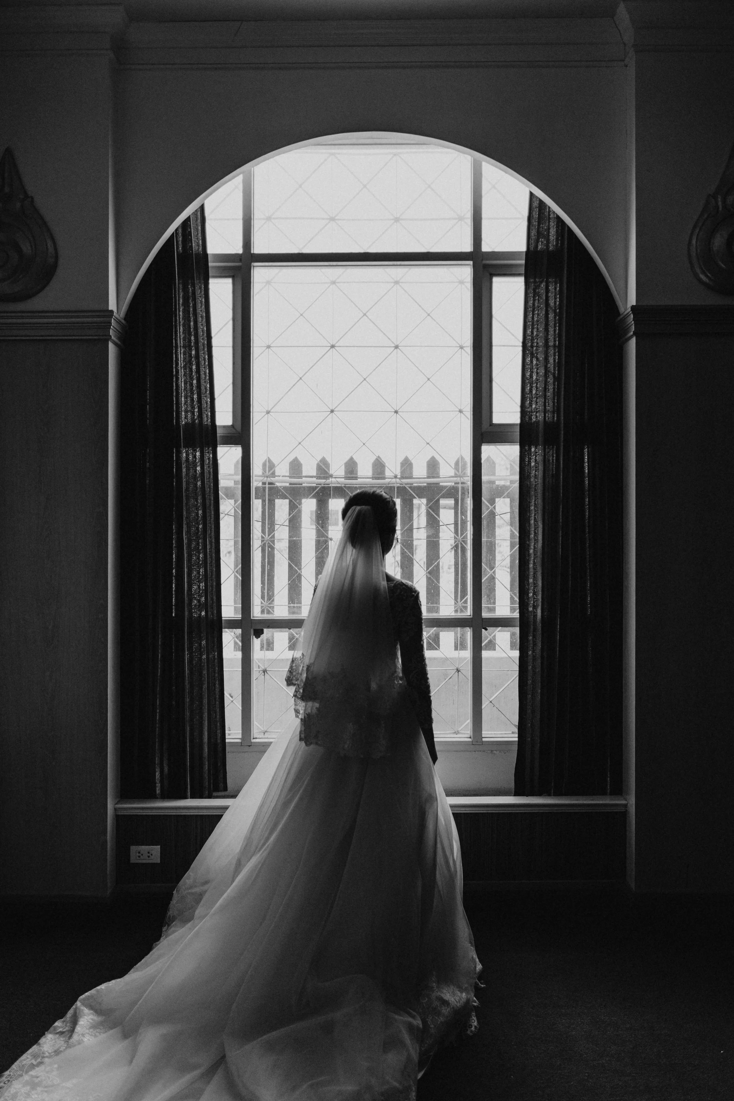 A bride in a wedding dress and veil standing in front of a large window with decorative panes and curtains, looking outside.