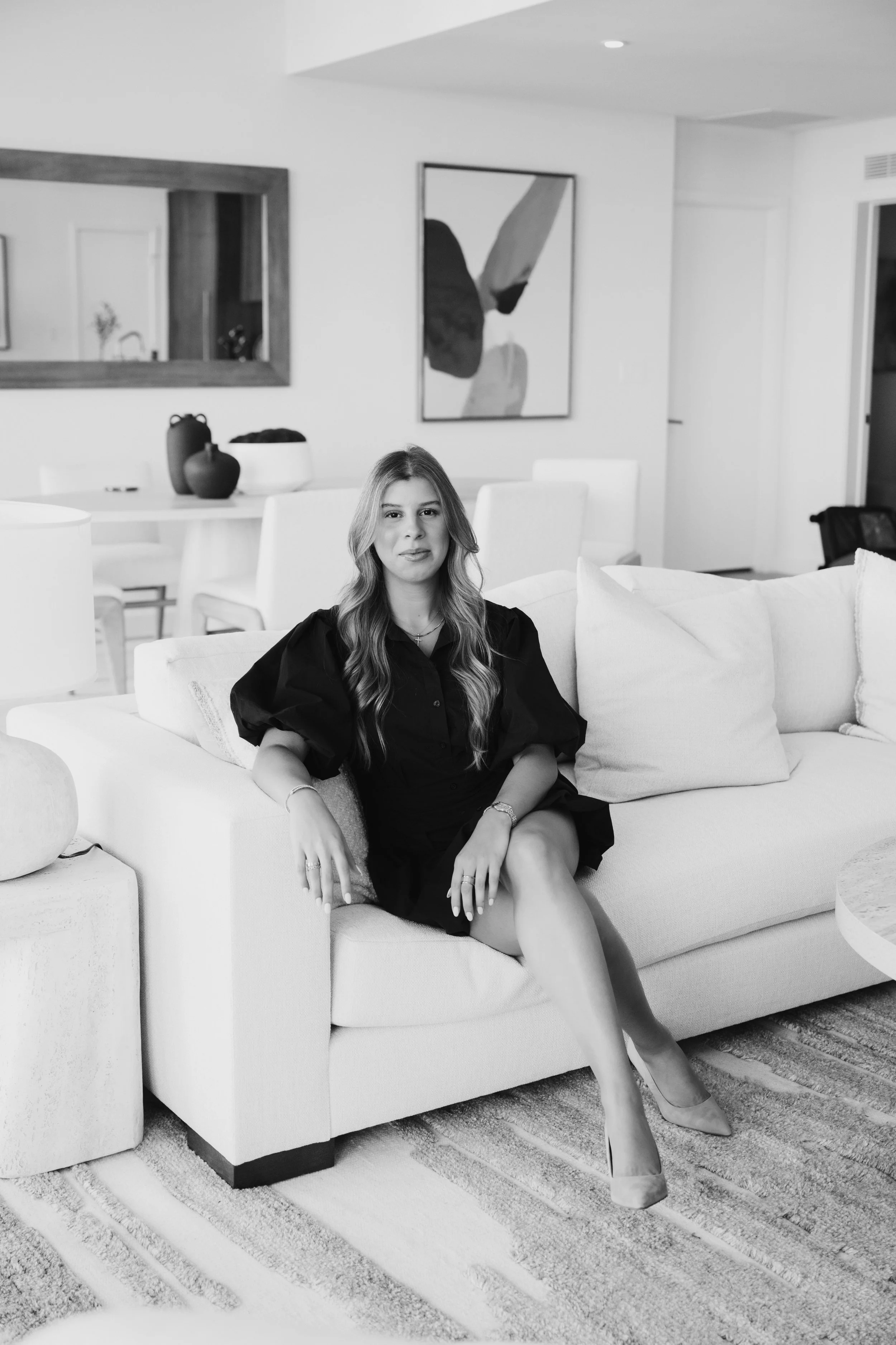 A woman with wavy hair sitting on a light-colored couch in a modern living room, wearing a dark dress and high heels, with legs crossed and hands resting on her lap.