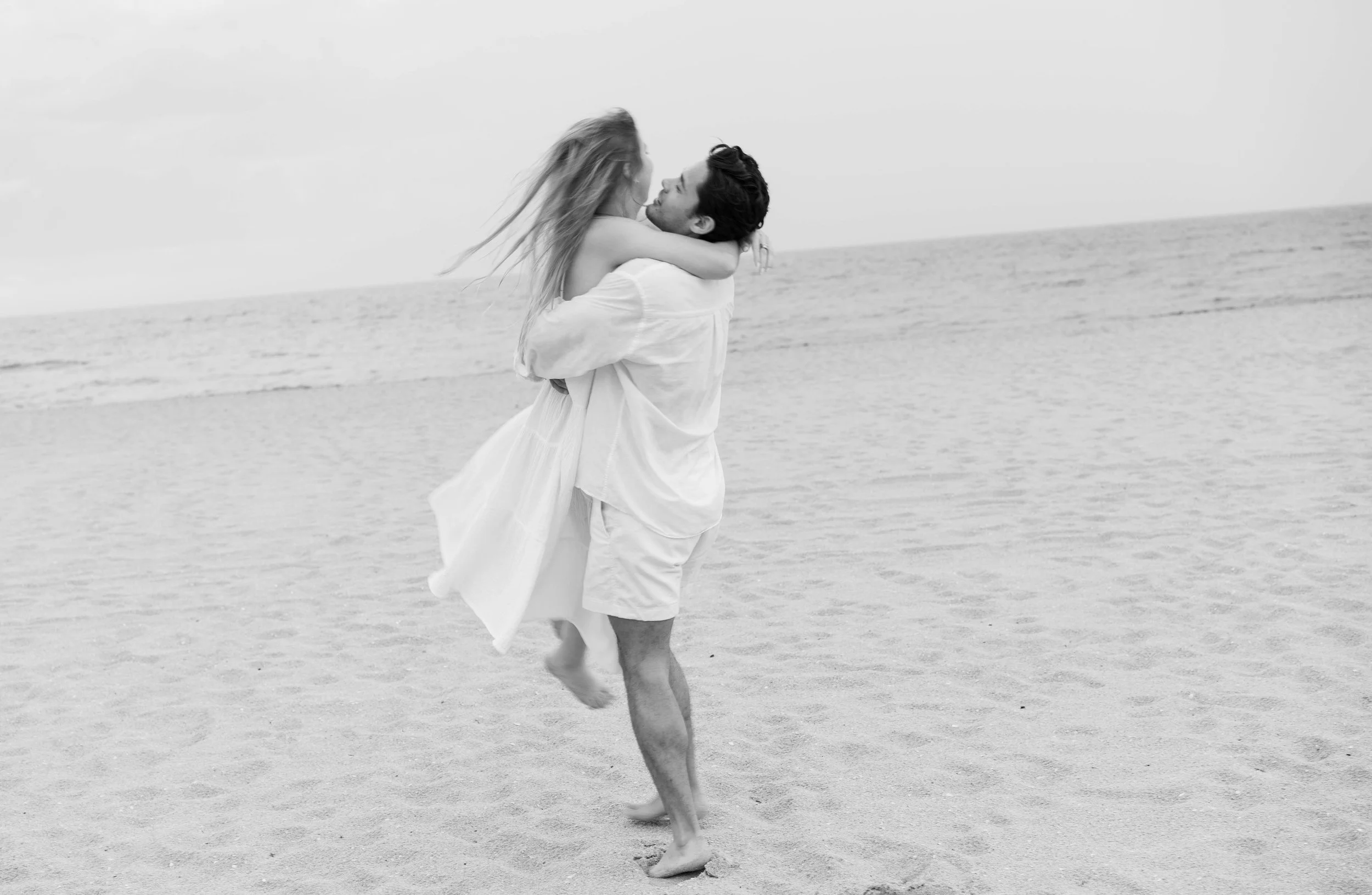 A man and woman on the beach are embracing, with the woman lifted in the man's arms. The woman has long hair and is wearing a flowing dress, while the man is in shorts and a shirt. The scene is in black and white, with the ocean and sky in the background.