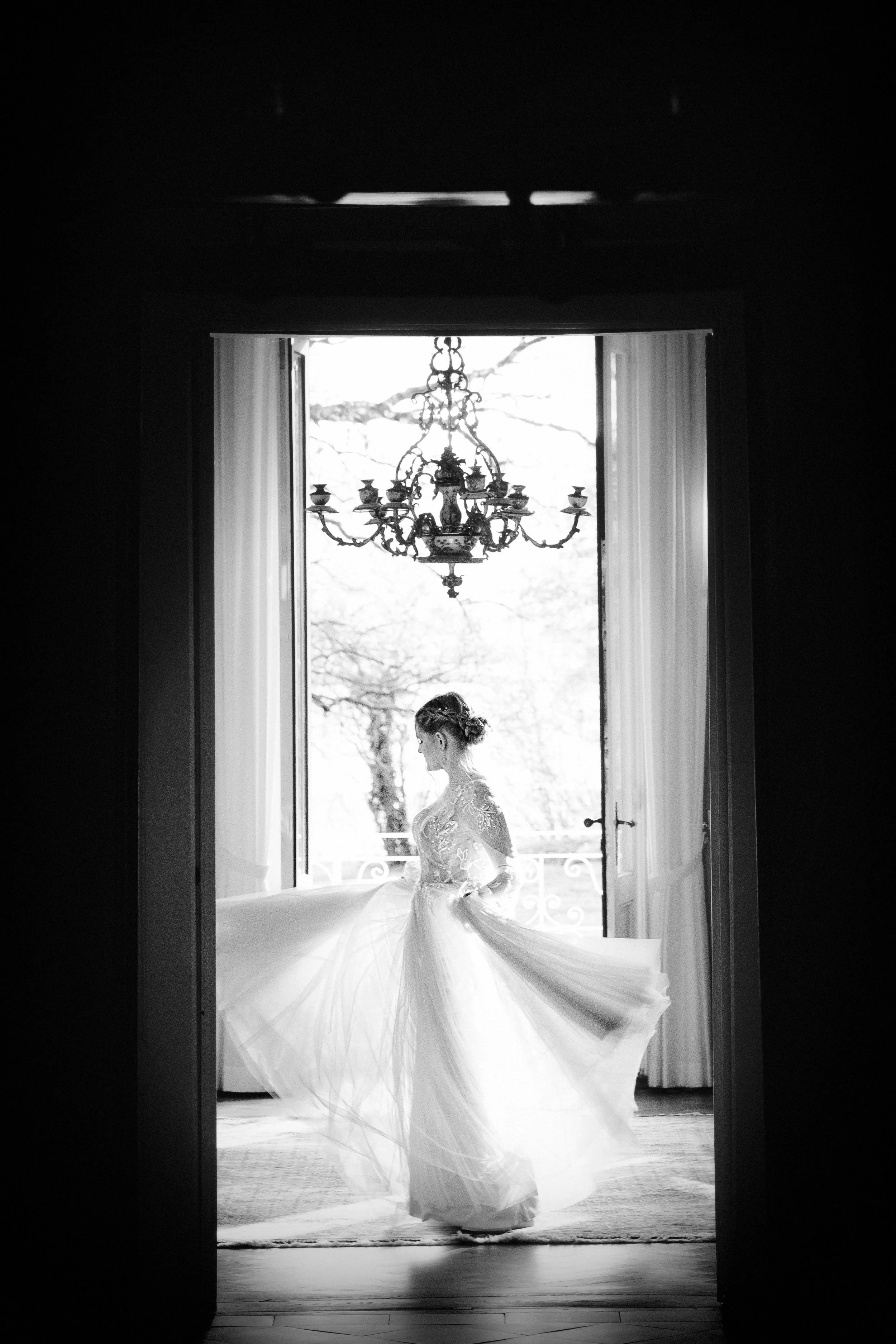 Black and white photo of a woman wearing a wedding dress standing in a doorway with open windows and a chandelier above, outdoor trees visible in the background.