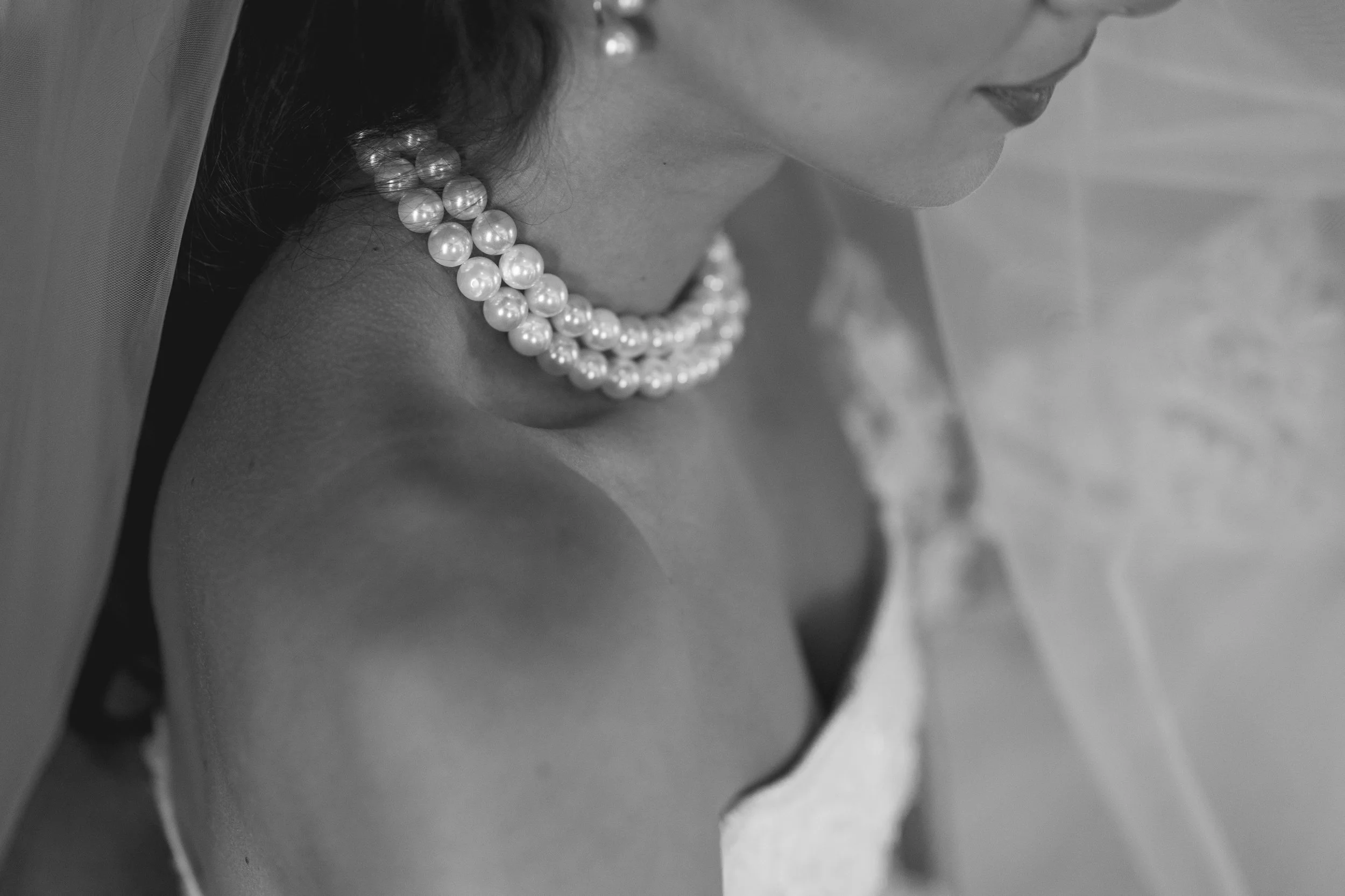 A close-up black and white photograph of a woman's neck and lower face, featuring multiple strands of pearl necklaces and a white veil.