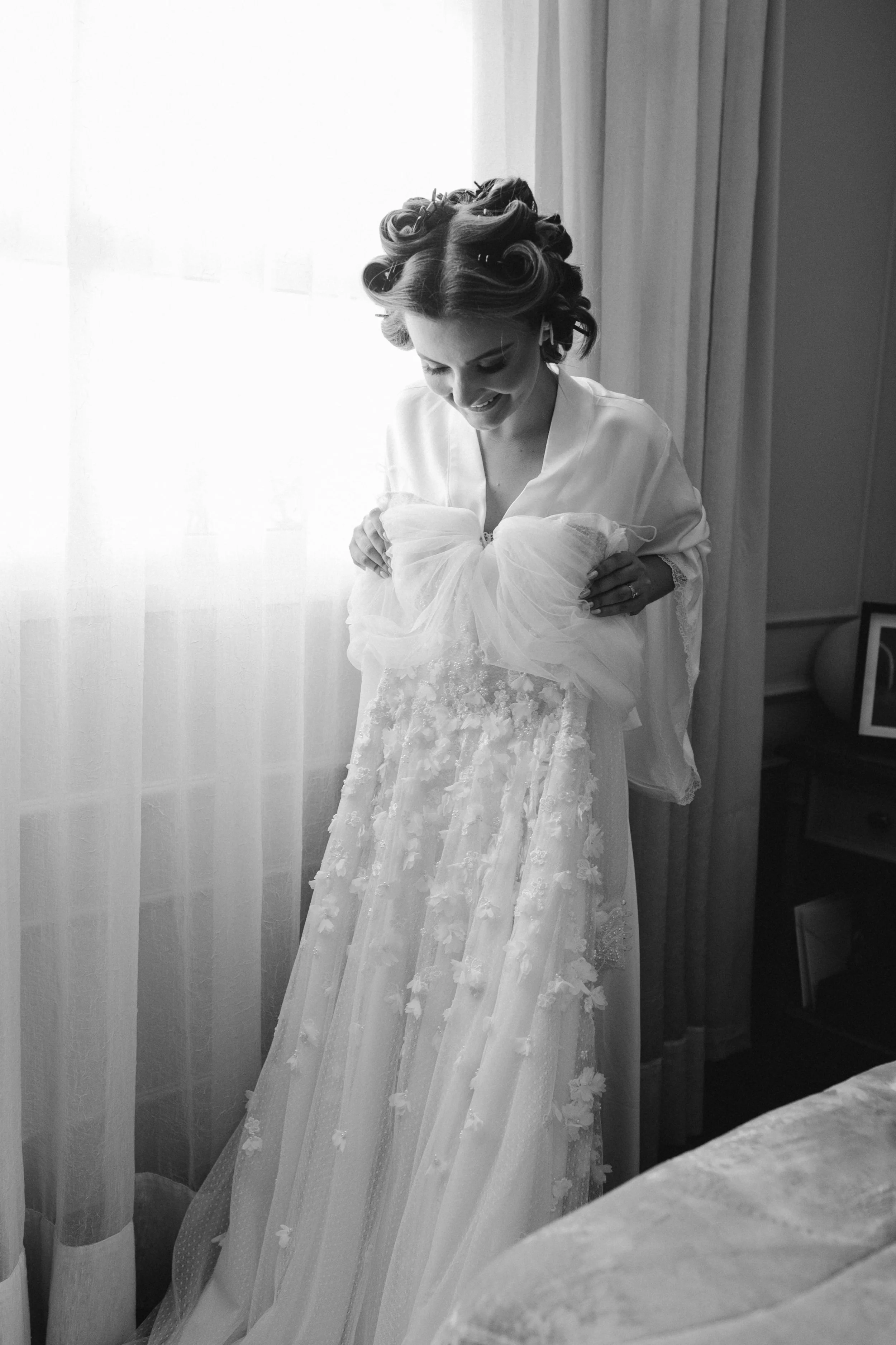 A woman with styled hair smiling while looking down, in a wedding dress holding a veil, standing indoors near a window with sheer curtains.