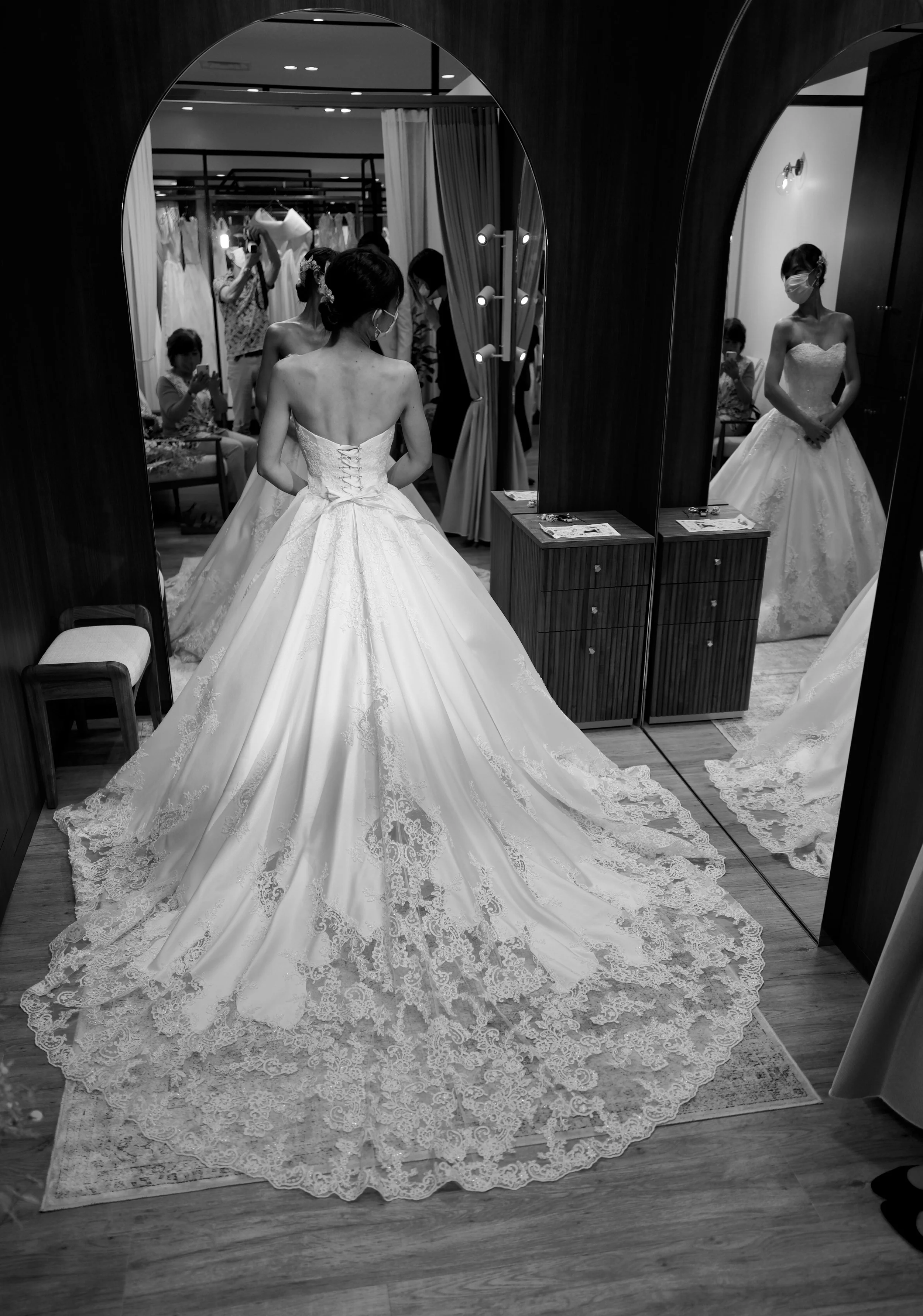 A woman trying on a wedding dress in front of a large mirror, with other people and hanging wedding dresses in the background.