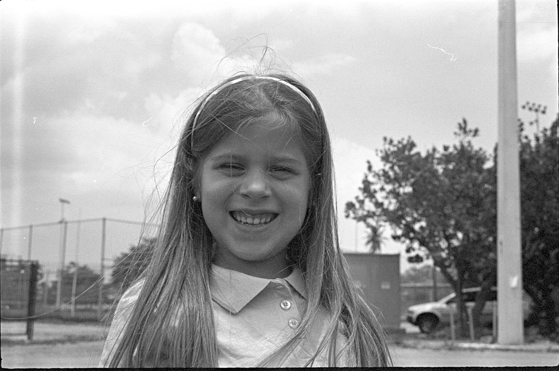 Black and white photo of a young girl with long hair, smiling with her eyes closed, outdoors near a fence, trees, and a parked car.