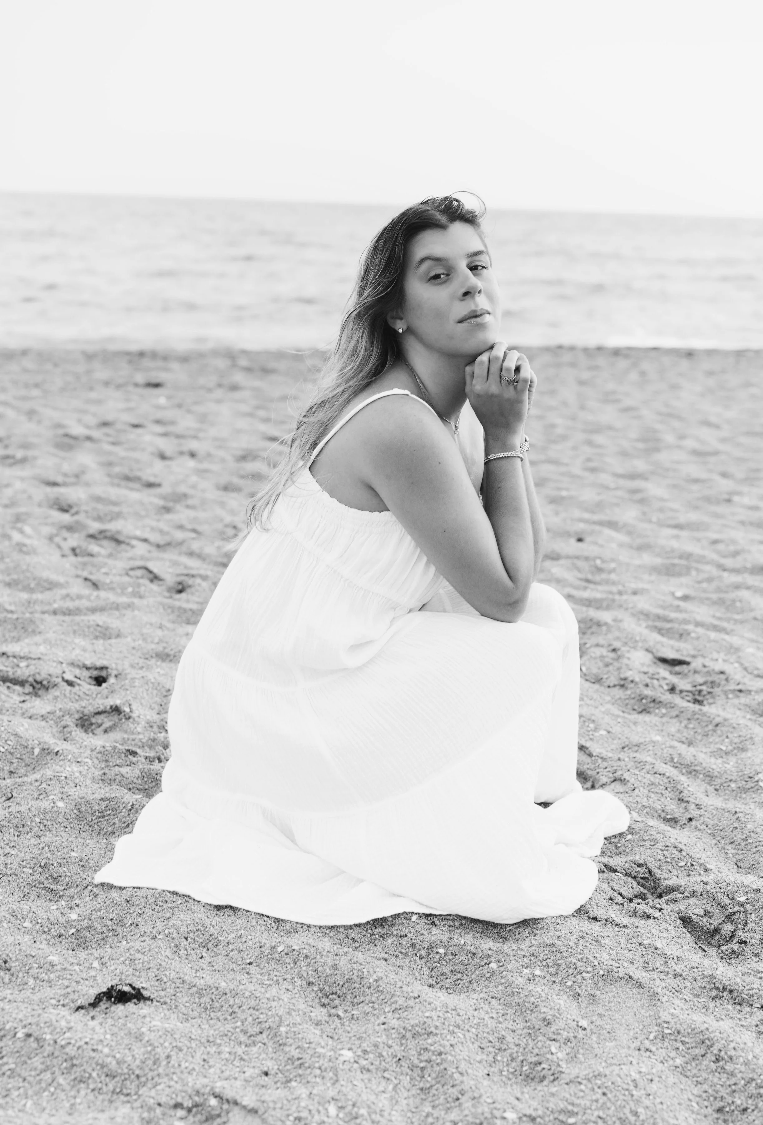 A woman sitting on the sandy beach in a white dress, with her chin resting on her hand, looking at the camera, with the ocean in the background.