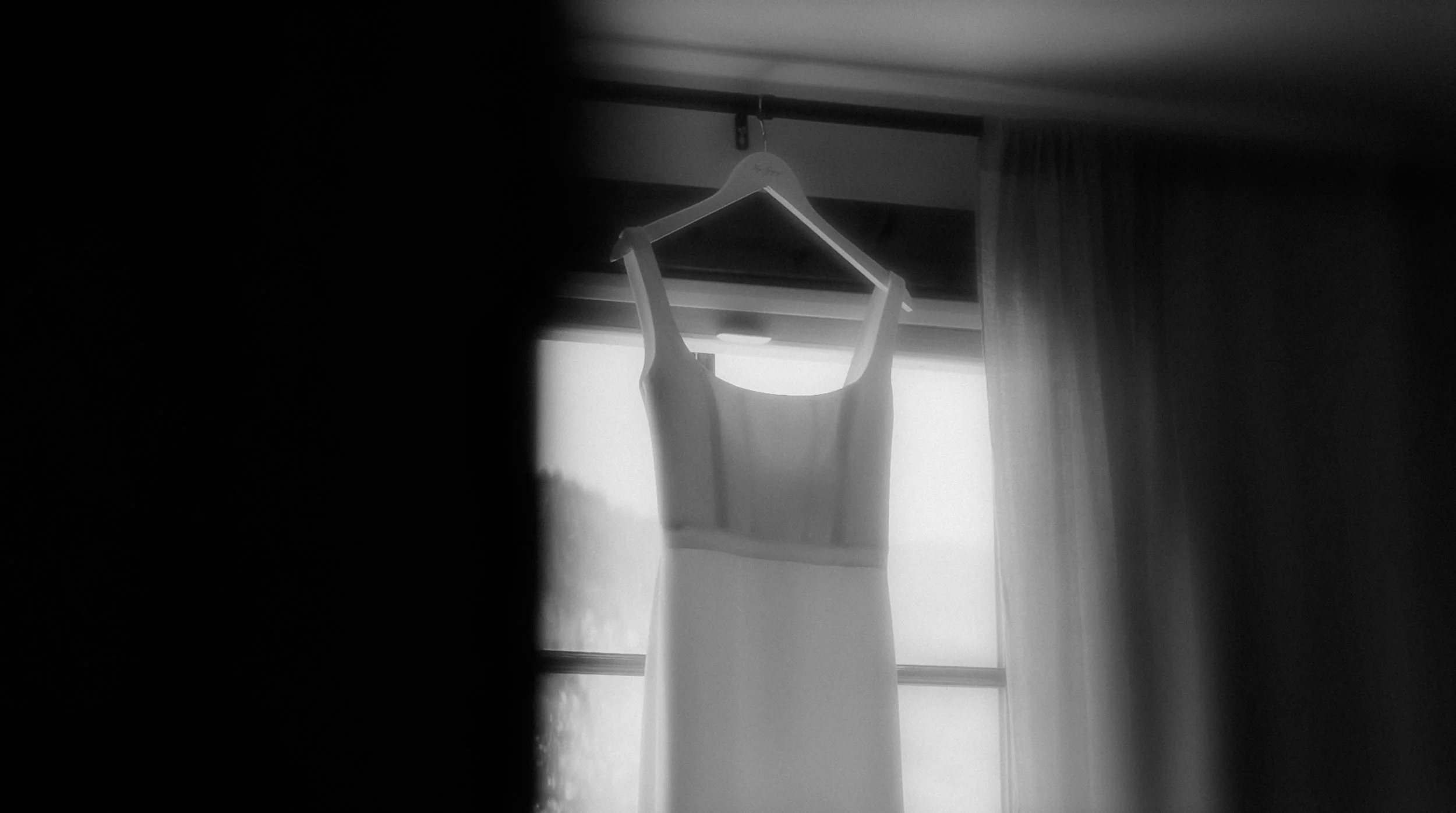 A white sleeveless dress hanging on a wooden hanger on a window, with curtains partially visible on the right side of the image. The photo is in black and white.
