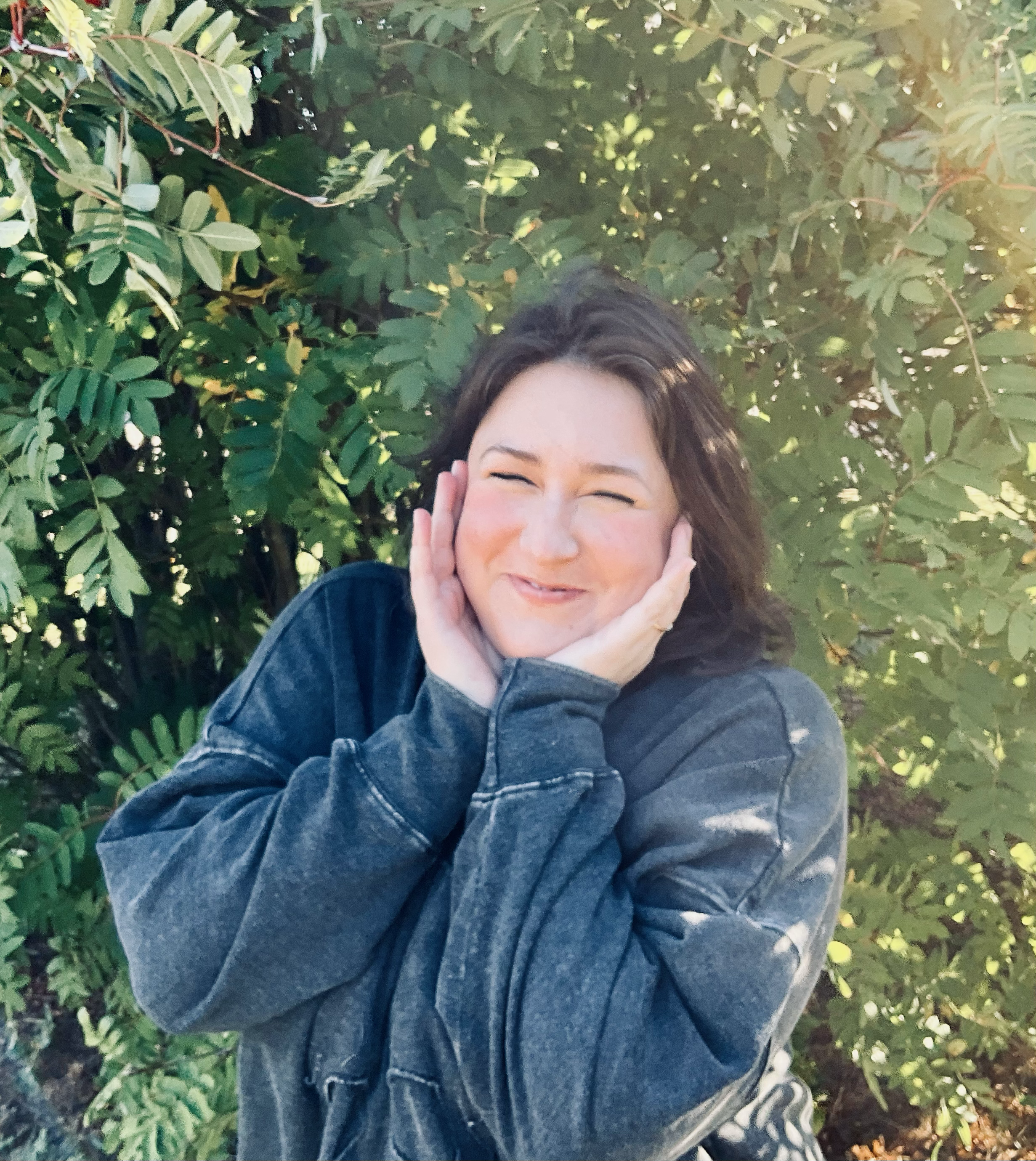 A woman smiling and covering her ears with her hands, standing outdoors near green leafy bushes or trees.