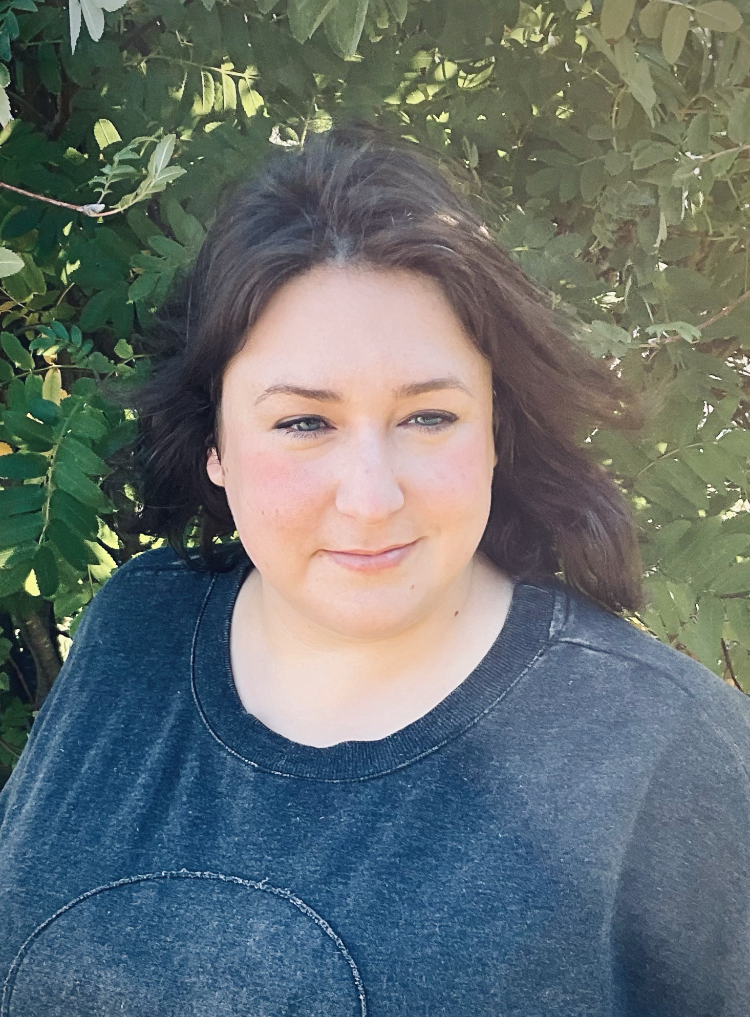 A woman with dark hair and blue eyes taking a selfie outdoors with green foliage in the background, wearing a dark gray shirt.