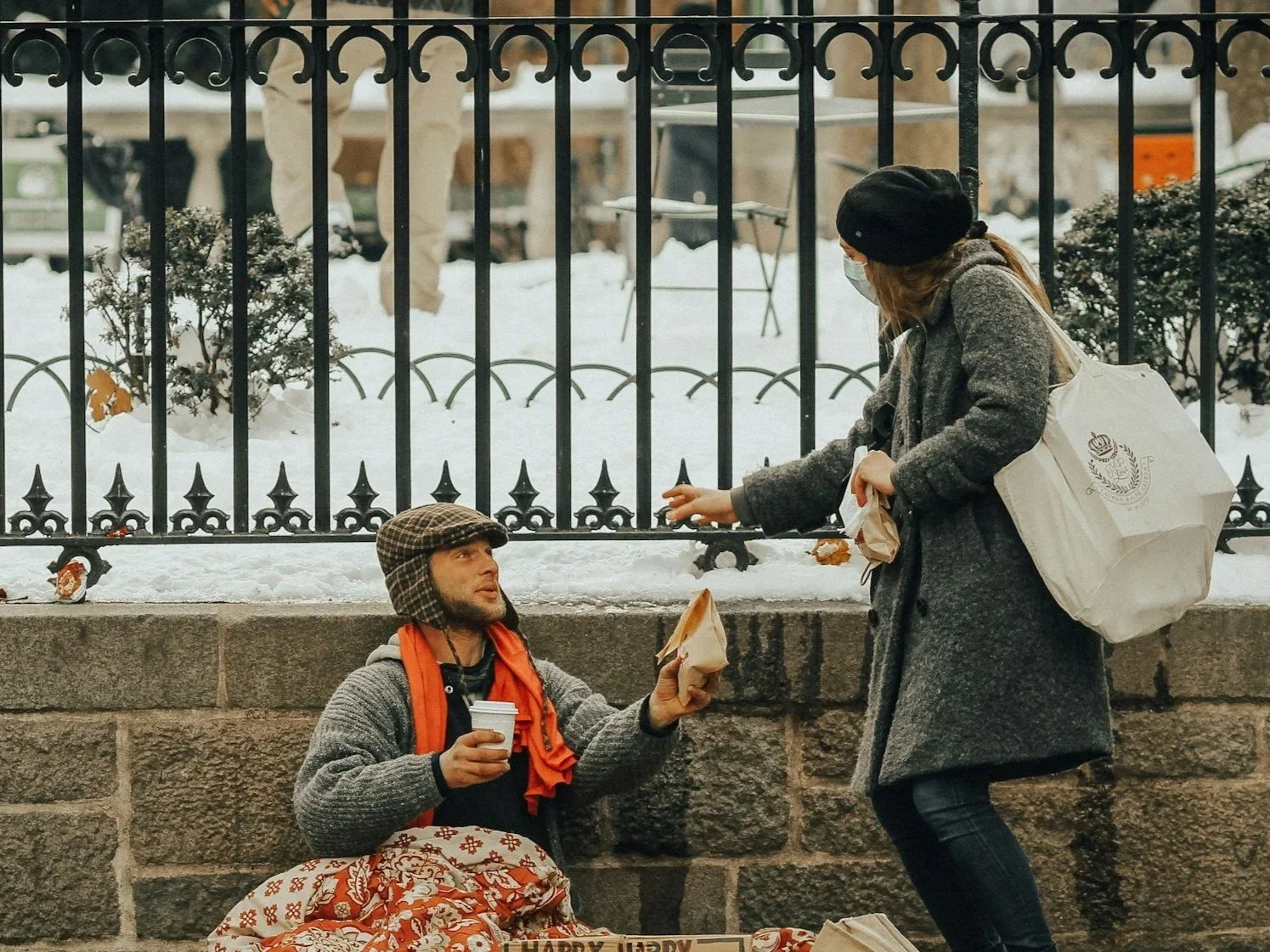 A woman with a backpack and face mask offers food to a homeless man sitting on a sidewalk against a stone wall. The man, wearing a plaid hat and gray sweater, reaches out for the food. Snow is visible behind a black iron fence.