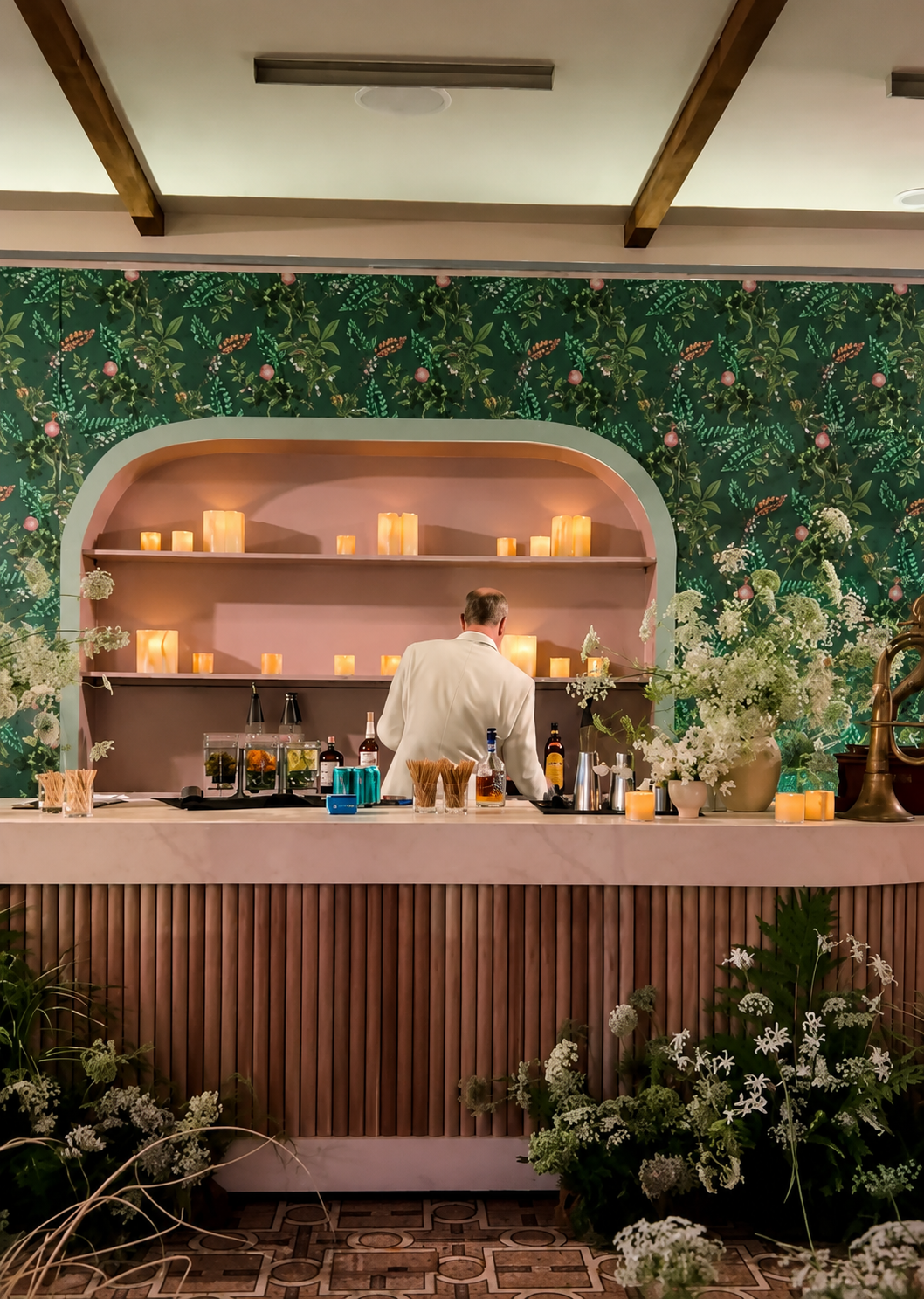 A bartender in a white suit preparing drinks at a decorated bar with candles, flowers, and a floral wallpaper background.